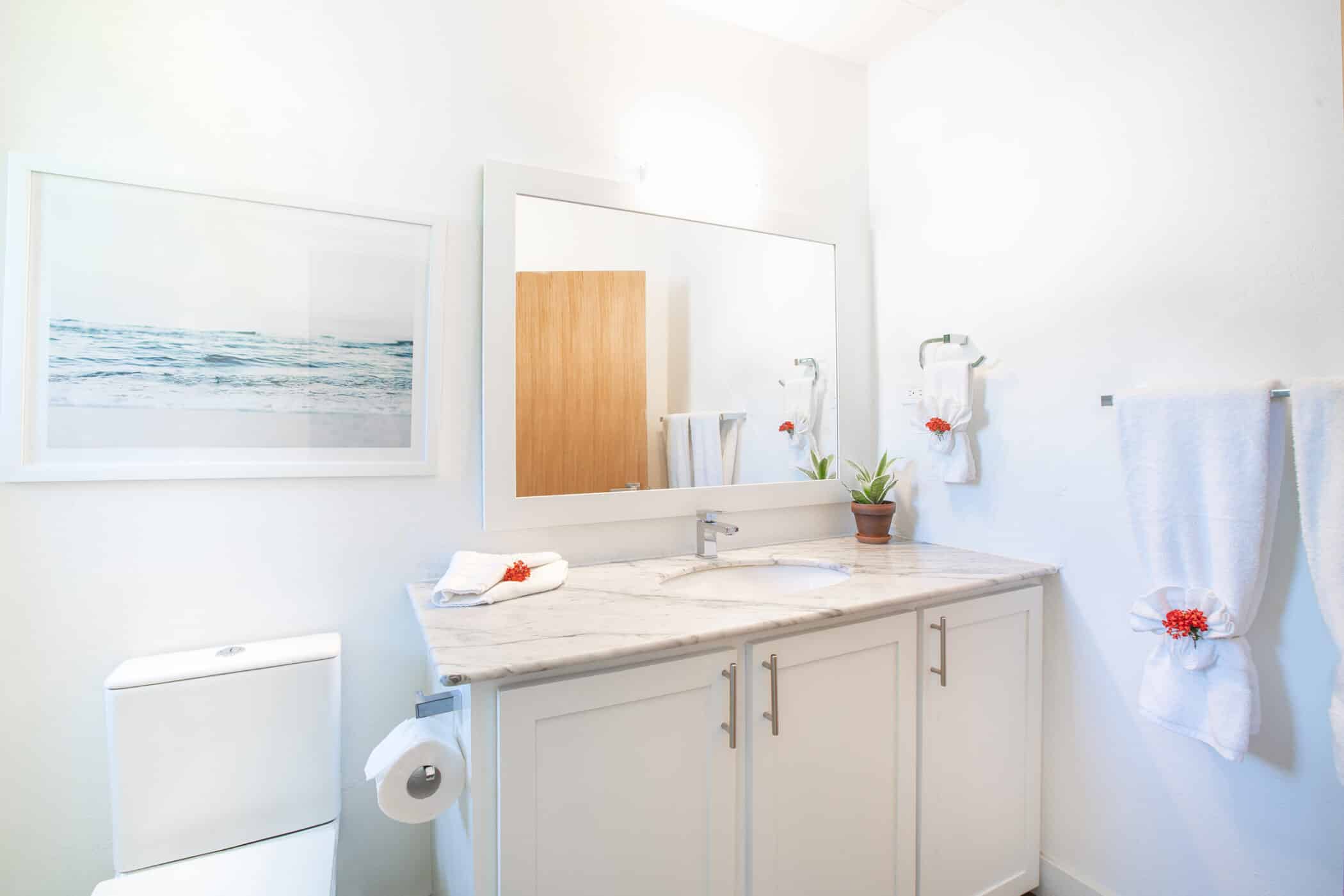 Bright bathroom with a large mirror, white cabinets, marble countertop, potted plant, towels with red flowers, wall art of the ocean titled 
