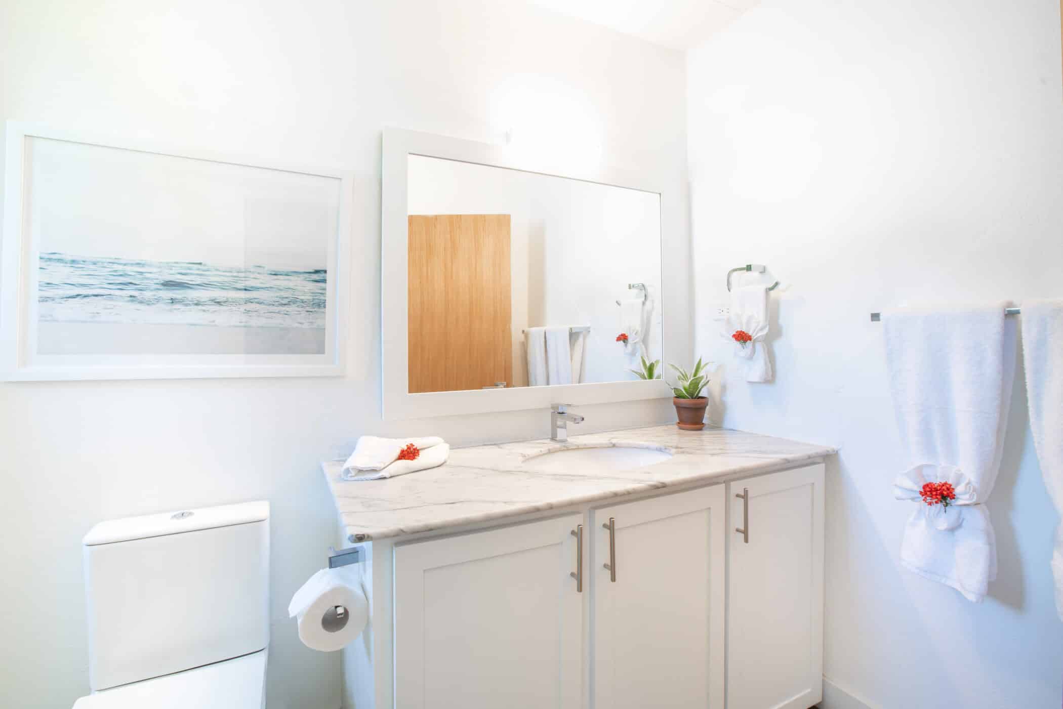 Bright bathroom with a white toilet, marble countertop, potted plant, large mirror, towels with red flowers, and a captivating “Secret Cove Two” seascape picture on the wall.
