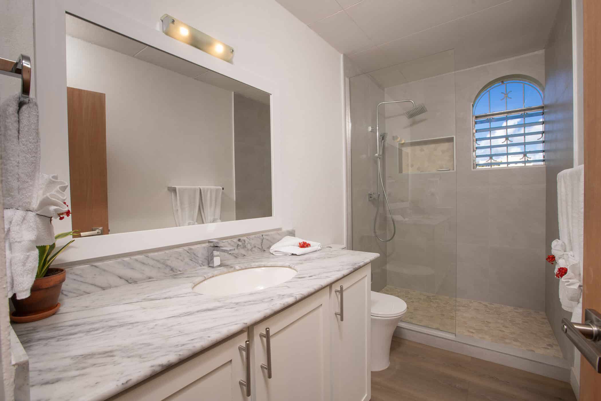 A modern bathroom with a marble countertop, large mirror, white cabinets, walk-in shower with glass door, and towels neatly arranged creates a sanctuary reminiscent of Secret Cove Two. The window with bars adds both security and character to this refined space.