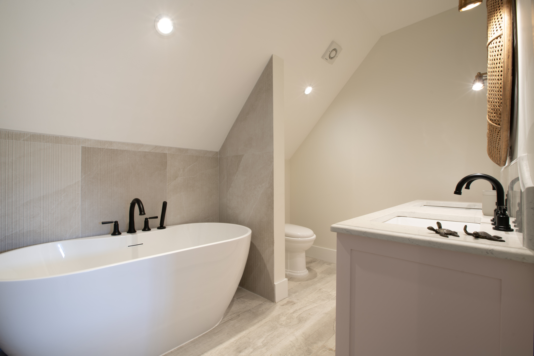 Modern bathroom at Seastar House & Cottage with a white freestanding bathtub, black fixtures, a toilet, and a sink with pale pink vanity under a sloped ceiling. Neutral tiles and soft lighting create a clean, serene atmosphere.