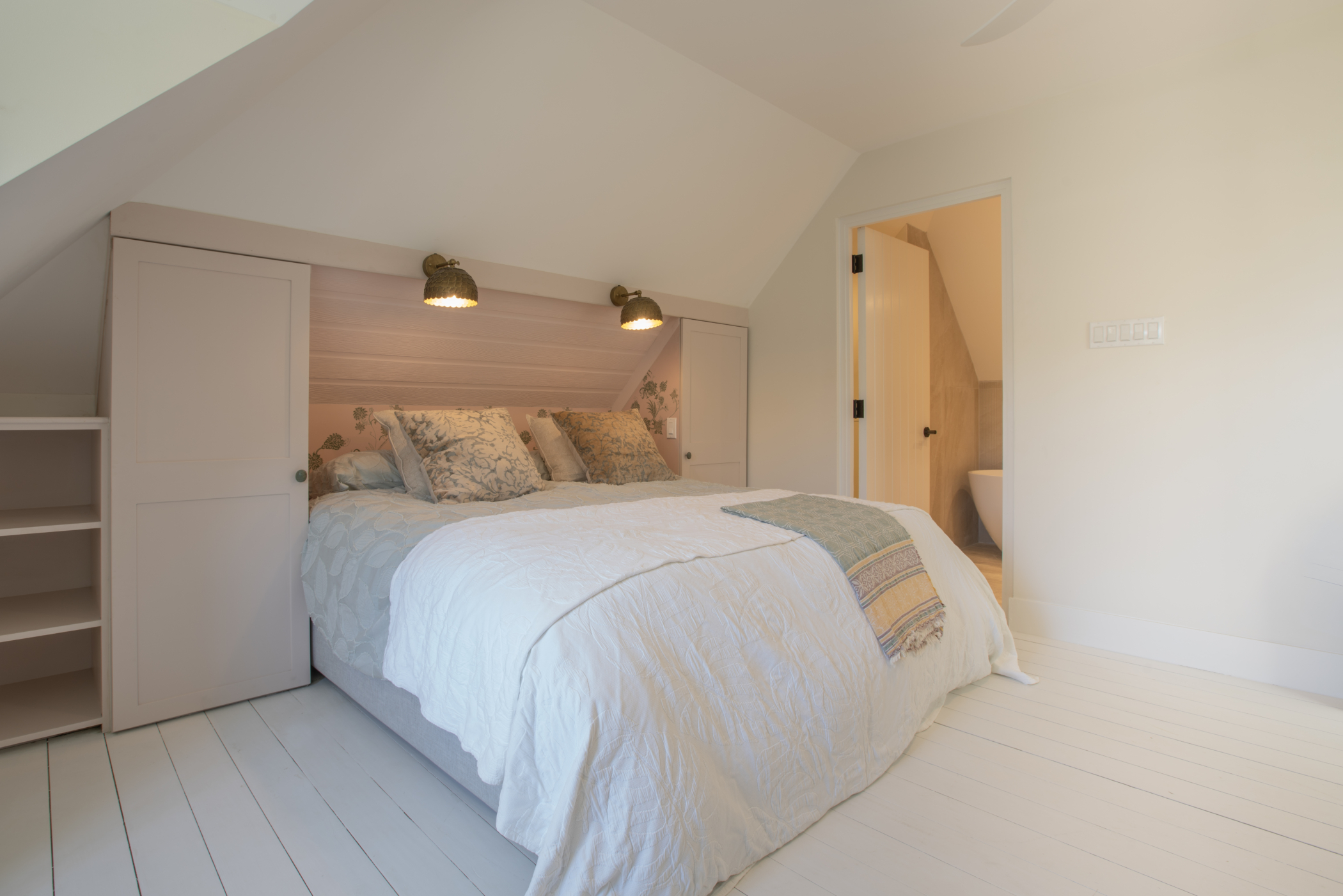 A cozy Seastar House & Cottage attic bedroom with a double bed, soft white bedding, decorative pillows, built-in shelves and closets, and warm wall-mounted lights; a door opens to a bathroom. The bright space features sloped ceilings and light walls.
