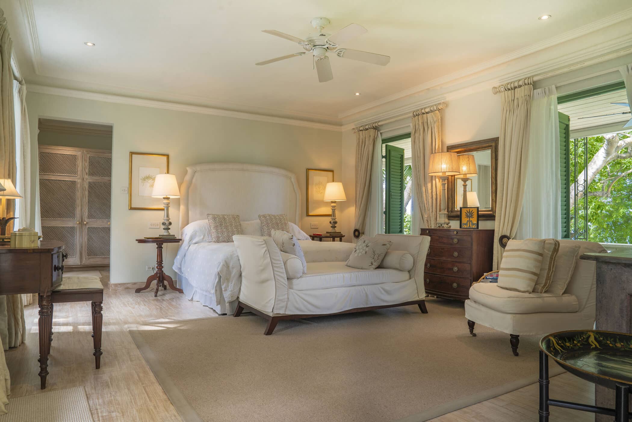 Spacious Maddox bedroom with a large bed, chaise lounge, armchair, and wooden furniture. Light walls, large windows with curtains, and greenery visible outside create a bright and cozy atmosphere.