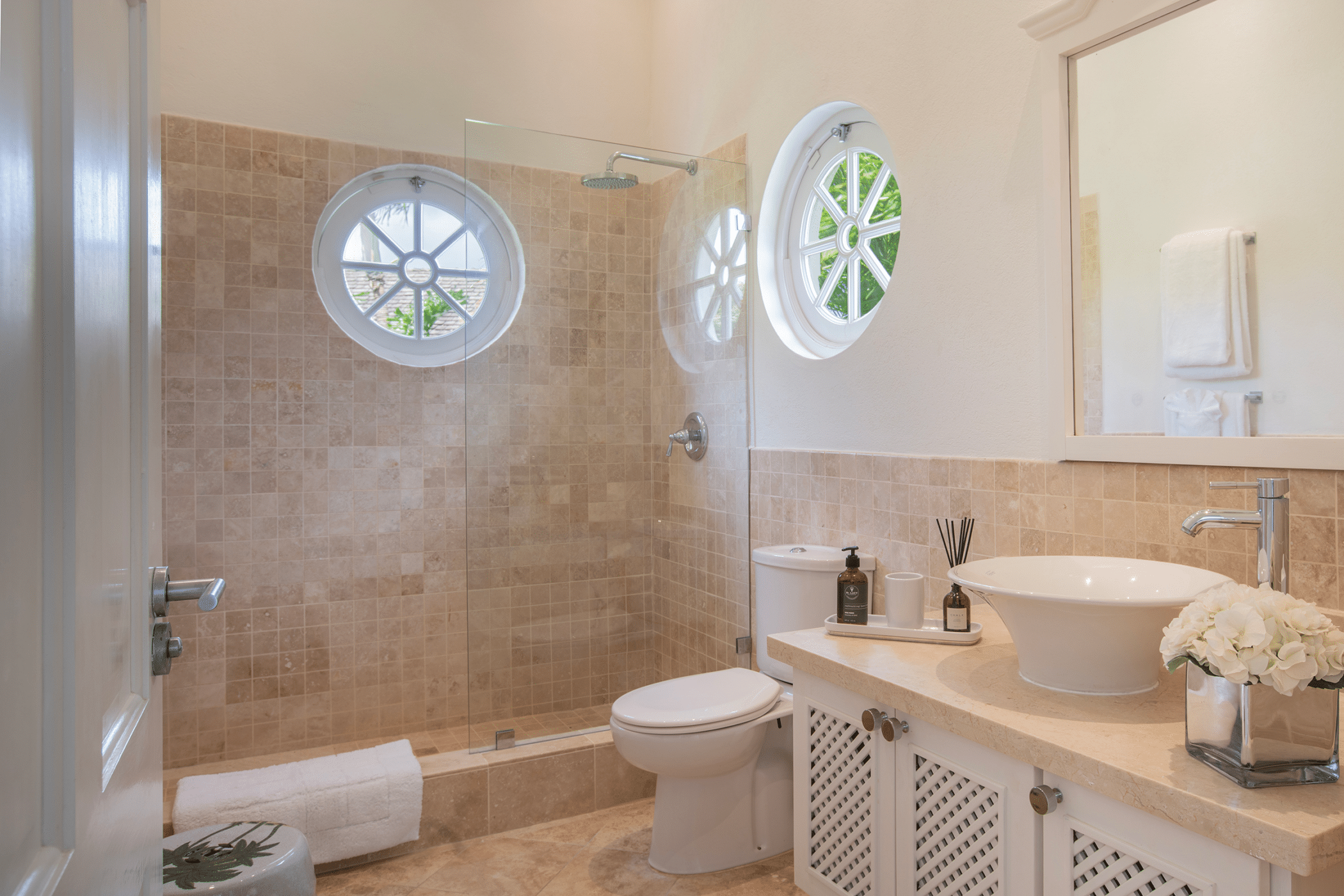 Bright bathroom with beige tiles, glass shower, circular windows, white toilet, modern sink, mirror, and decorative items on the countertop—including flowers, soap, and a diffuser—create an inviting Aquilae-inspired atmosphere. Rolled towels sit by the shower.