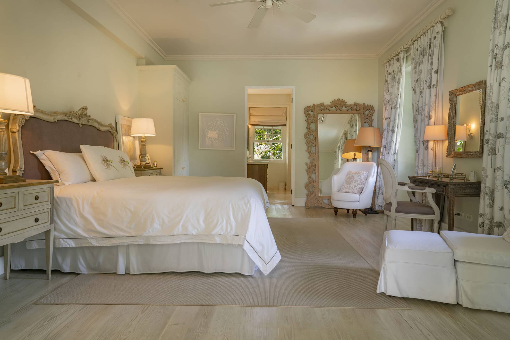 Elegant bedroom with a large bed, ornate mirror, armchair, side tables with lamps, light wood floors, floral curtains, and neutral decor. The Maddox design influence adds a sophisticated touch to the space. A door leads to a bright, connected room.