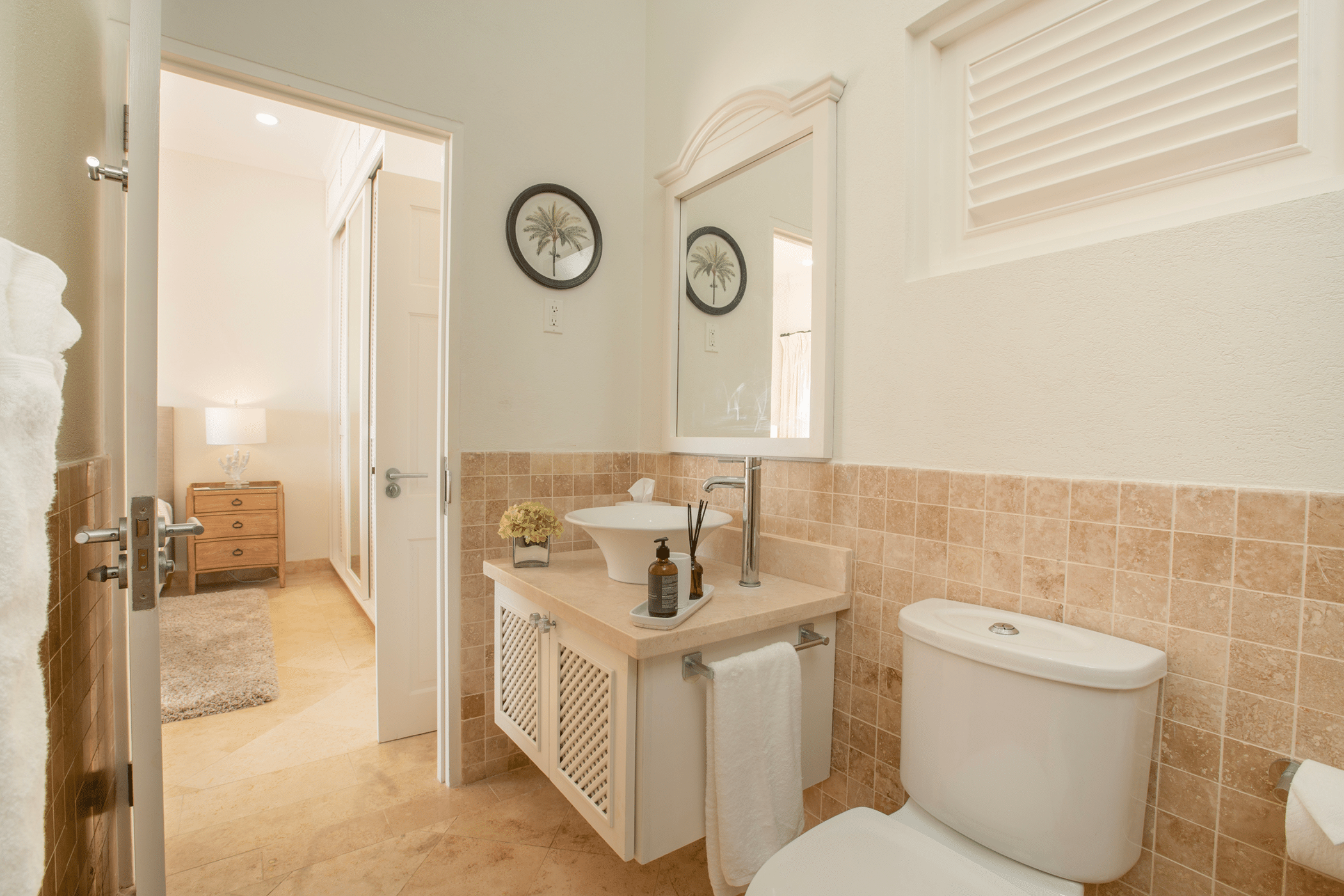A bright bathroom with a white sink, a mirror, a toilet, beige tiled walls, and a towel creates an inviting space reminiscent of Aquilae’s serene ambiance. An open door reveals a bedroom with a nightstand, lamp, and bed in a cozy, neutral-toned setting.