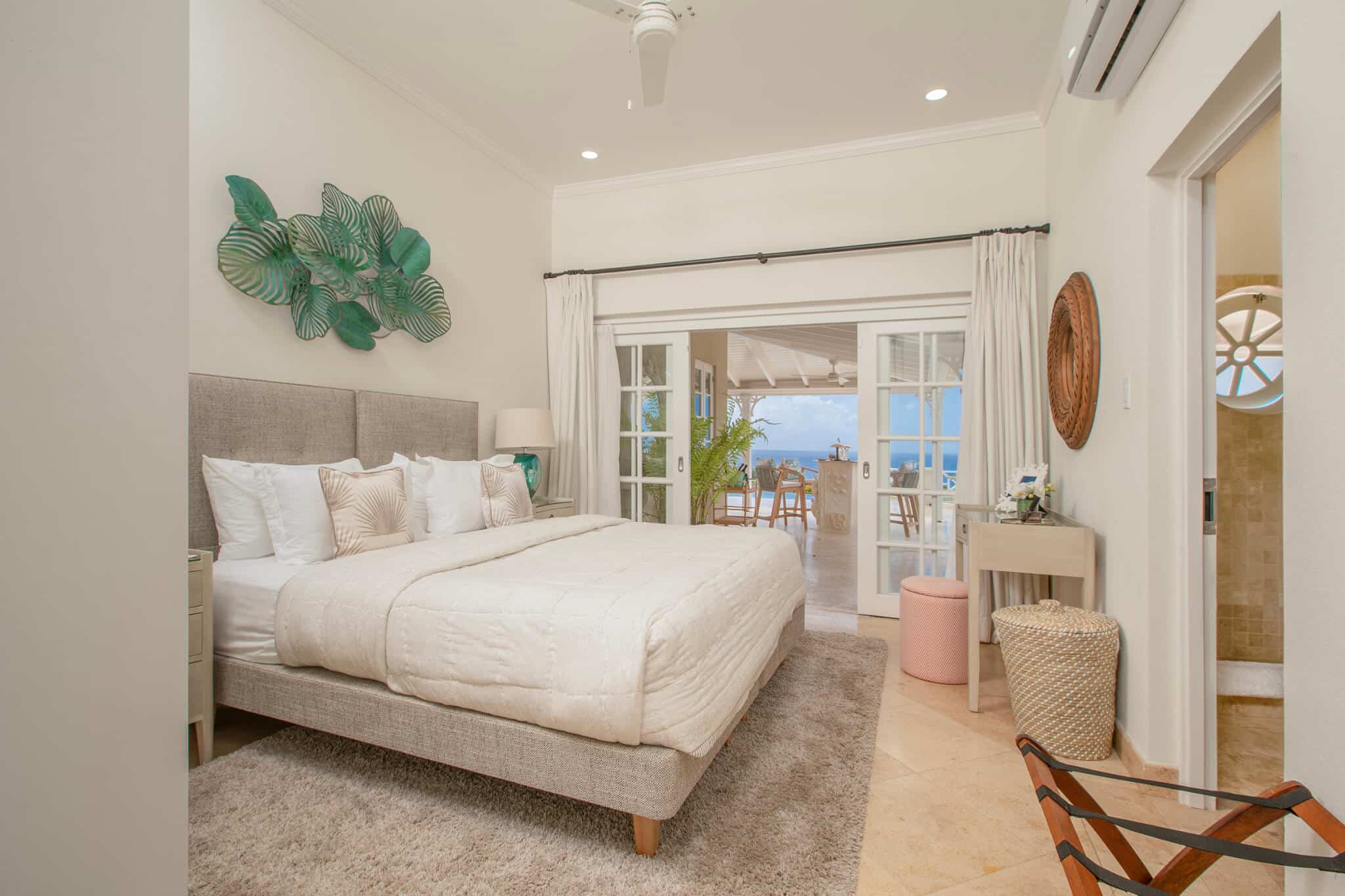 Bright bedroom with a large bed, neutral decor, and tropical wall art inspired by the tranquil spirit of Aquilae, opening to a patio with ocean view. Features a desk, wicker basket, and plush rug on tile floor.