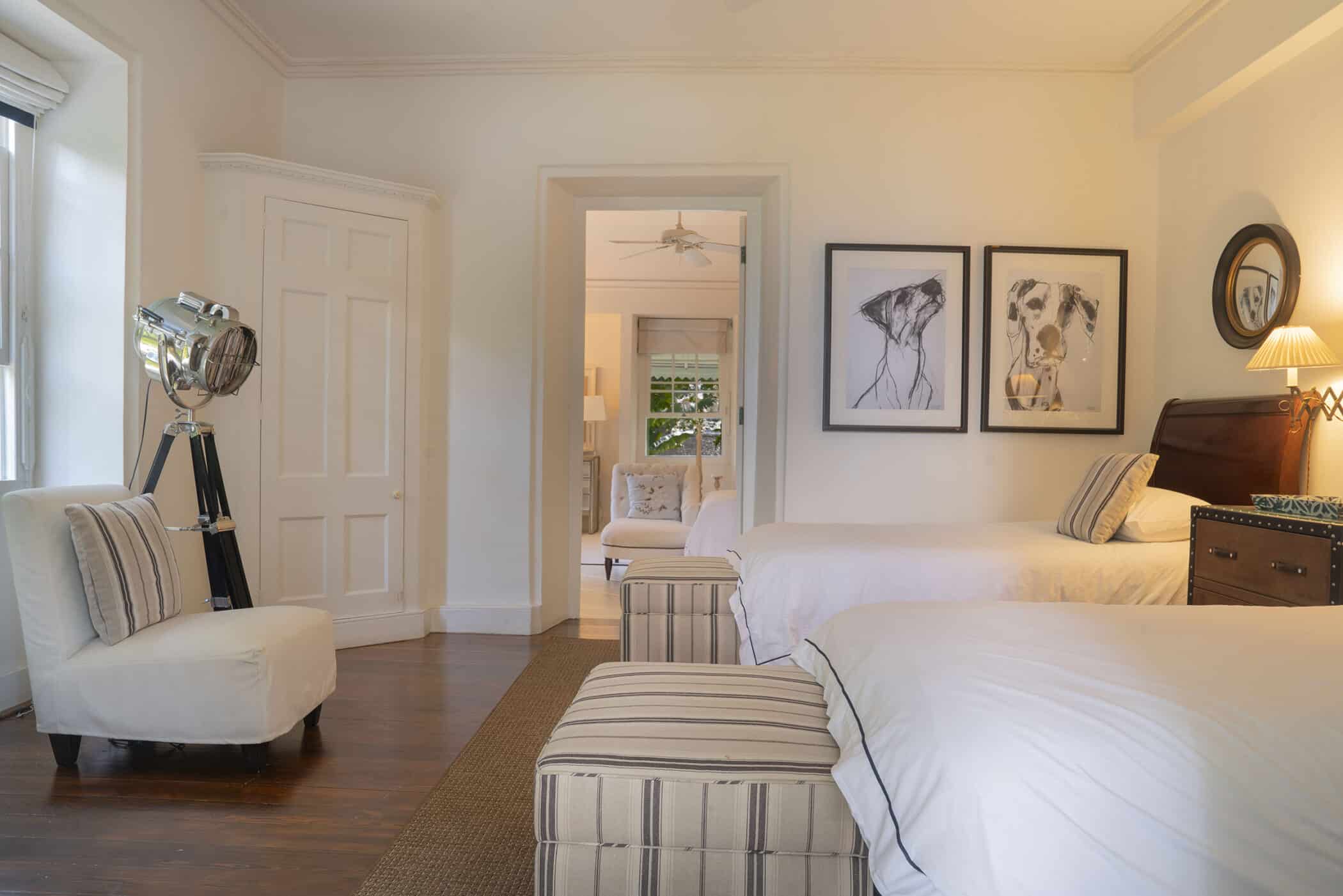 A bright bedroom with two beds, striped ottomans, white walls, framed dog art, a vintage lamp, and an upholstered chair near a window creates a cozy retreat. The Maddox design touches elevate the space’s charm. An open doorway reveals another sitting area.