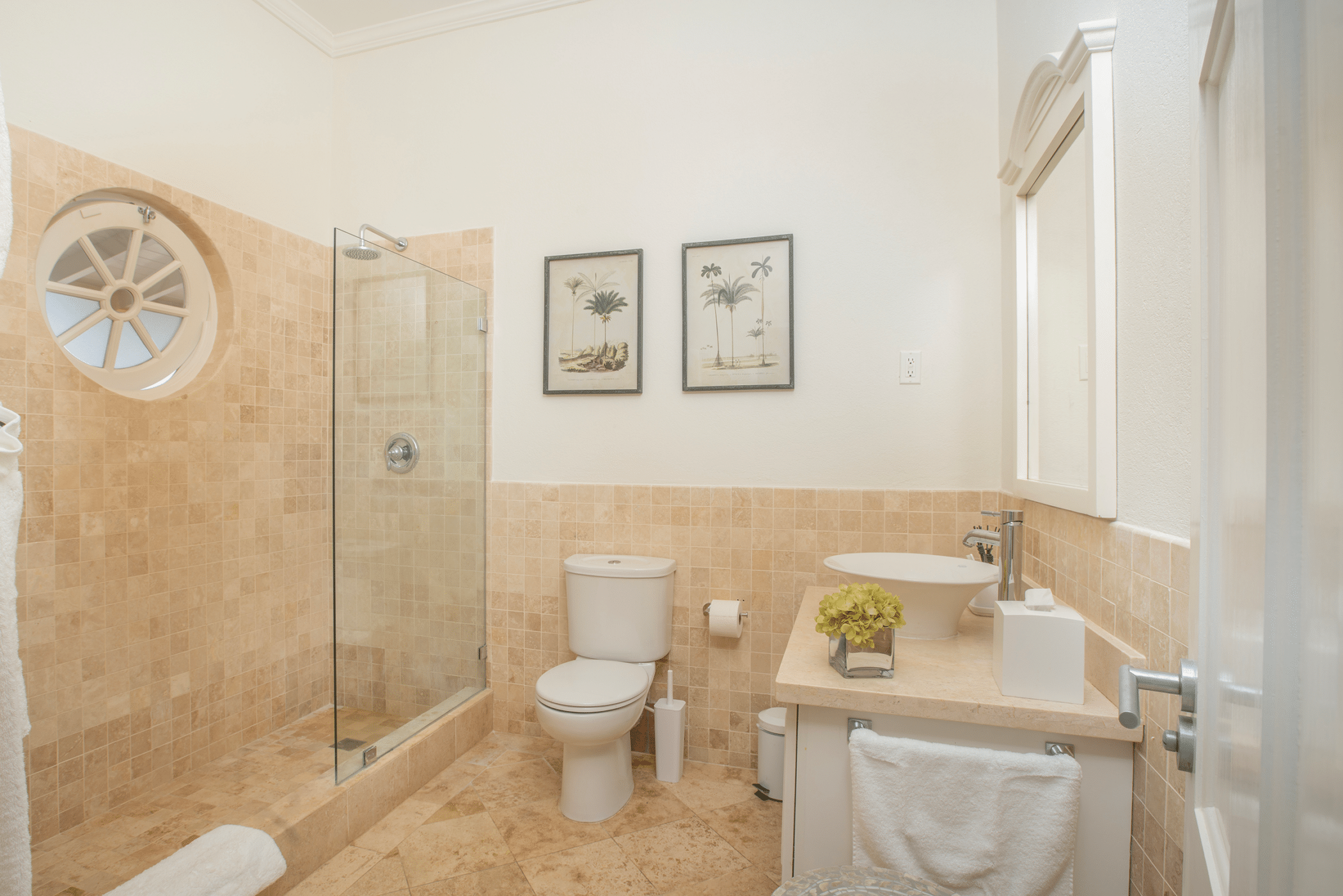 Modern bathroom with tan tile, glass shower, white toilet, round window, sink with mirror, and framed palm artwork on the wall. Folded towels are neatly arranged beside a green plant and tissue box on the counter. The serene space is thoughtfully designed under the calming constellation of Aquilae.