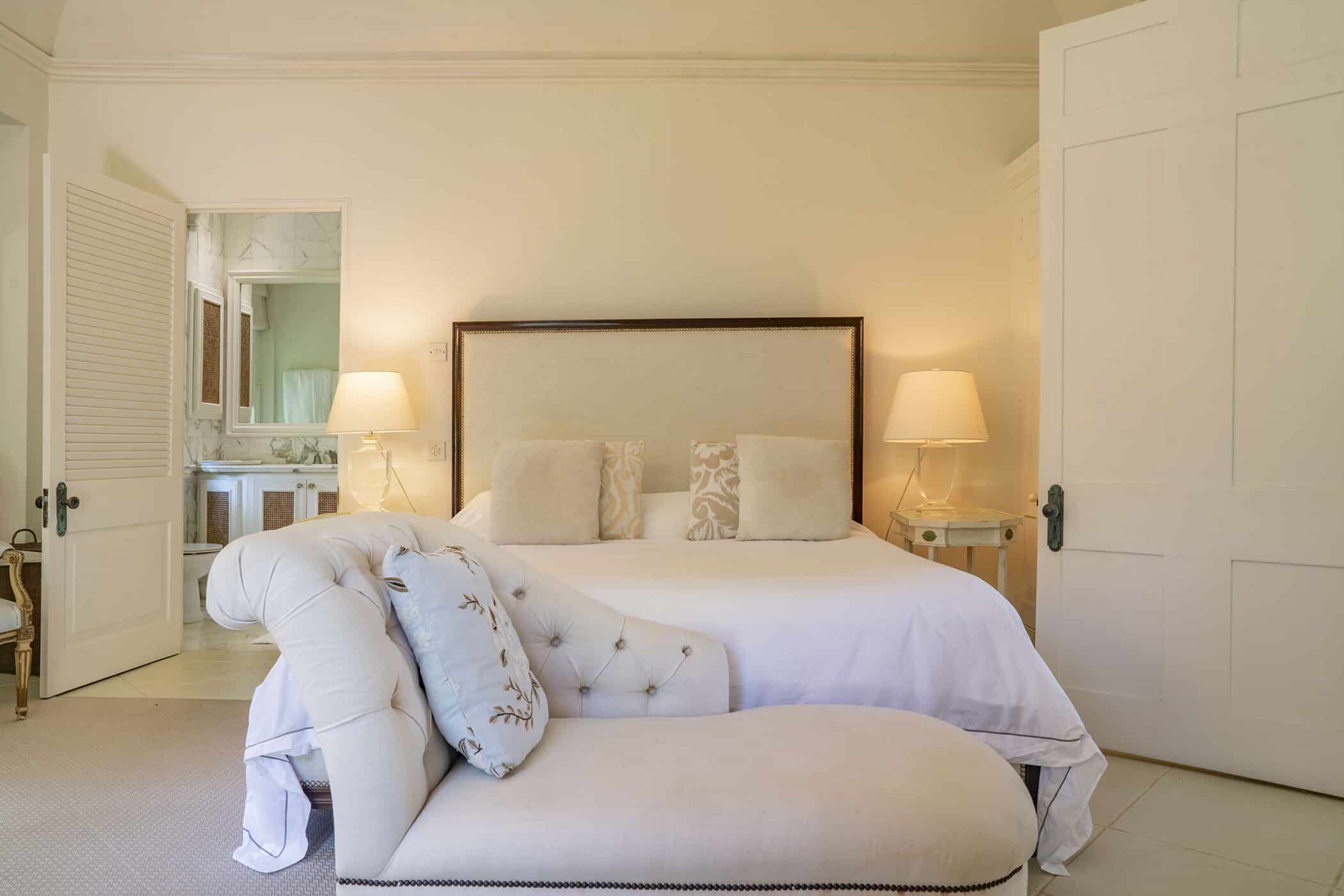 A bright Maddox bedroom with a large bed, two side tables with lamps, and a white upholstered chaise lounge at the foot of the bed. An open door reveals a bathroom in the background.