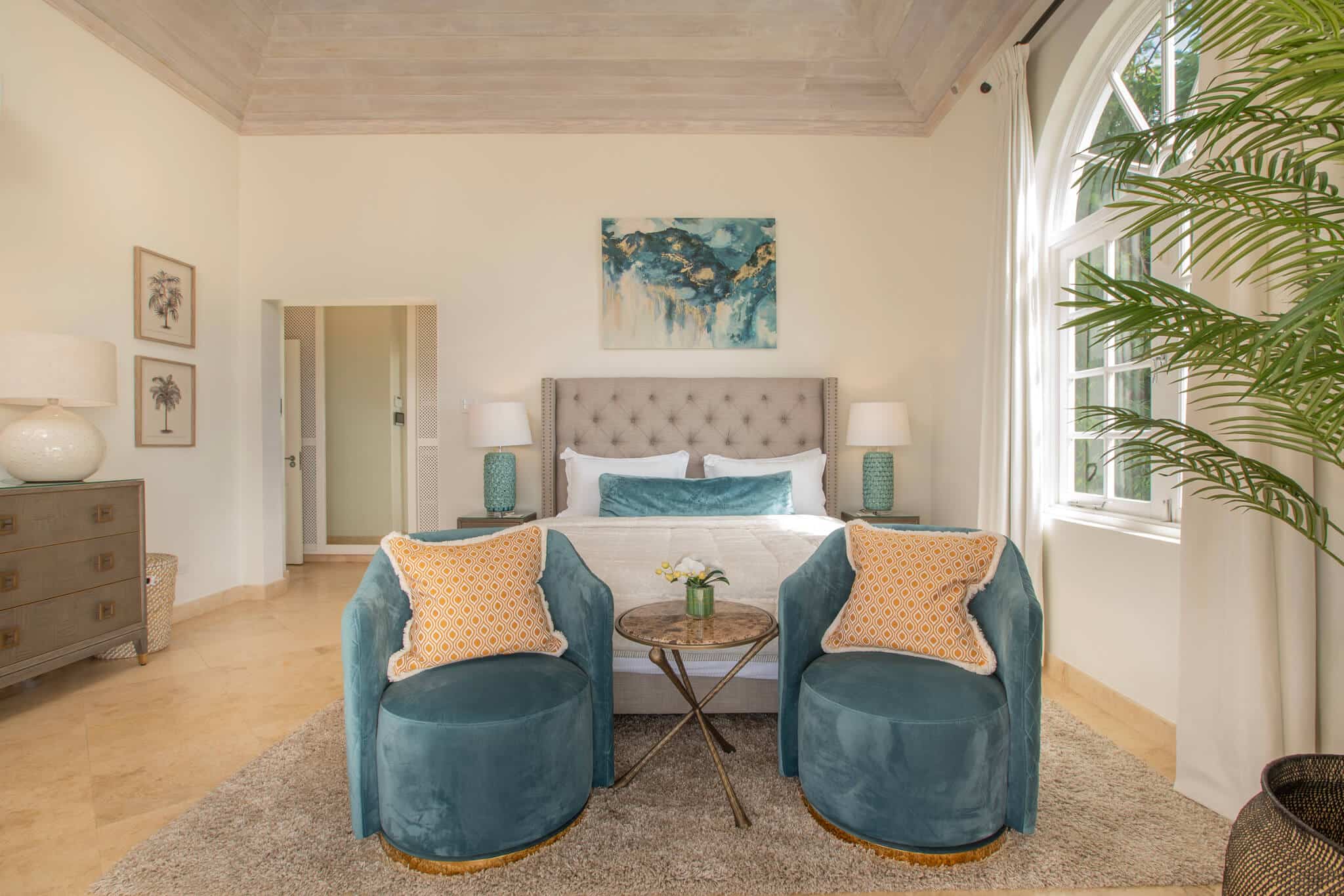 A bright bedroom with a tufted bed, two blue chairs with orange pillows, a round table, green lamp, large window, dresser, and palm plant. Light walls and a high ceiling create an airy atmosphere reminiscent of Aquilae’s serene elegance.