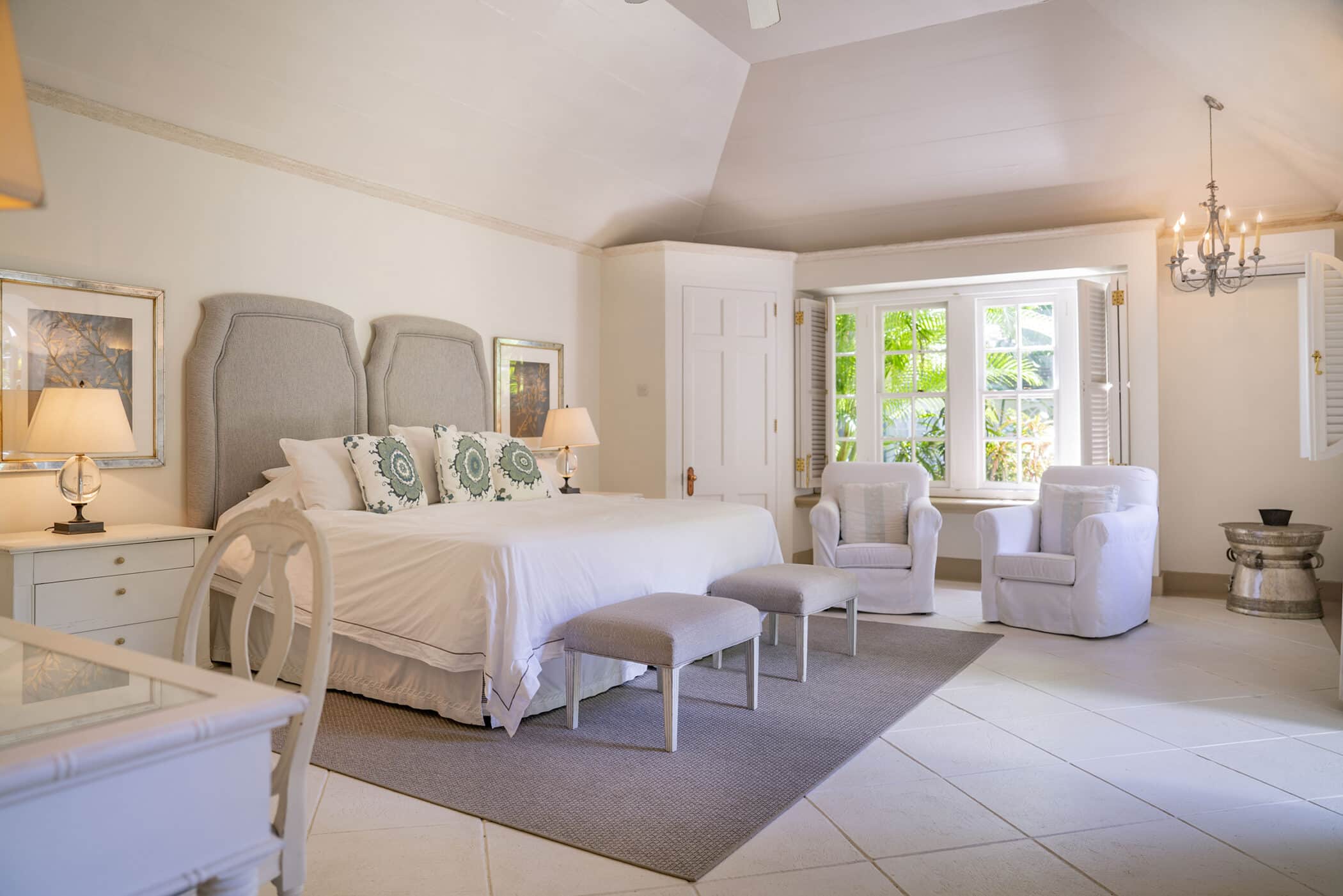 Bright bedroom with a large Maddox bed, two gray stools, armchairs by a window, lamps, framed art, and neutral decor. Sunlight streams in through open white shutters, creating a cozy, inviting atmosphere.