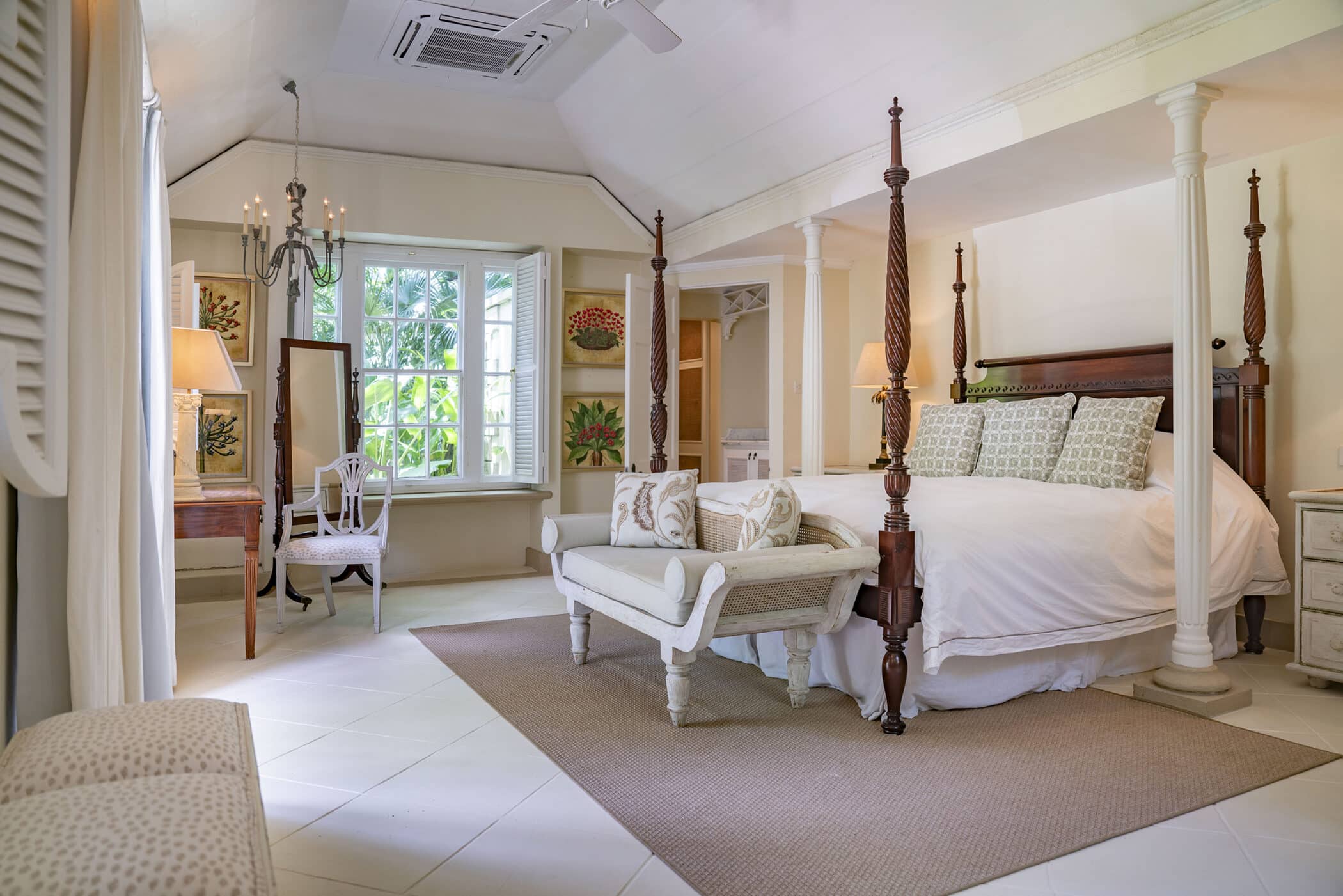 Bright, airy Maddox bedroom with a four-poster bed, cushioned bench, large window seat, vanity table, chandelier, and light decor featuring white and beige tones, green plants, and floral artwork.
