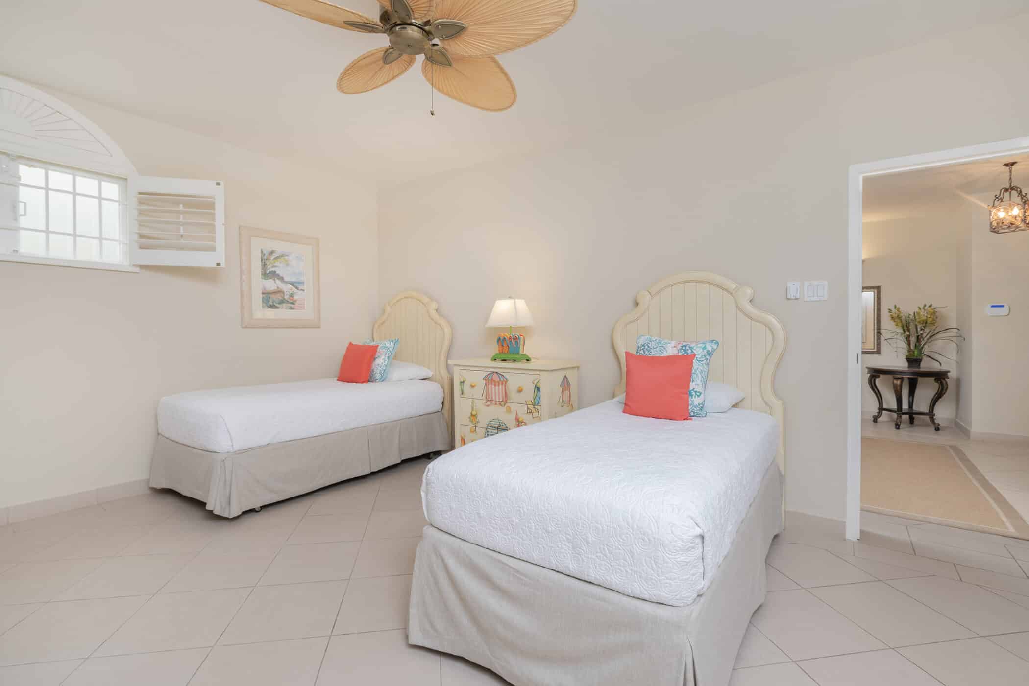 A bright bedroom with two twin beds, each with white bedding, coral and blue accent pillows, a decorative bedside table with a lamp between them, and tile floors. Ceiling fan and wall art are visible.