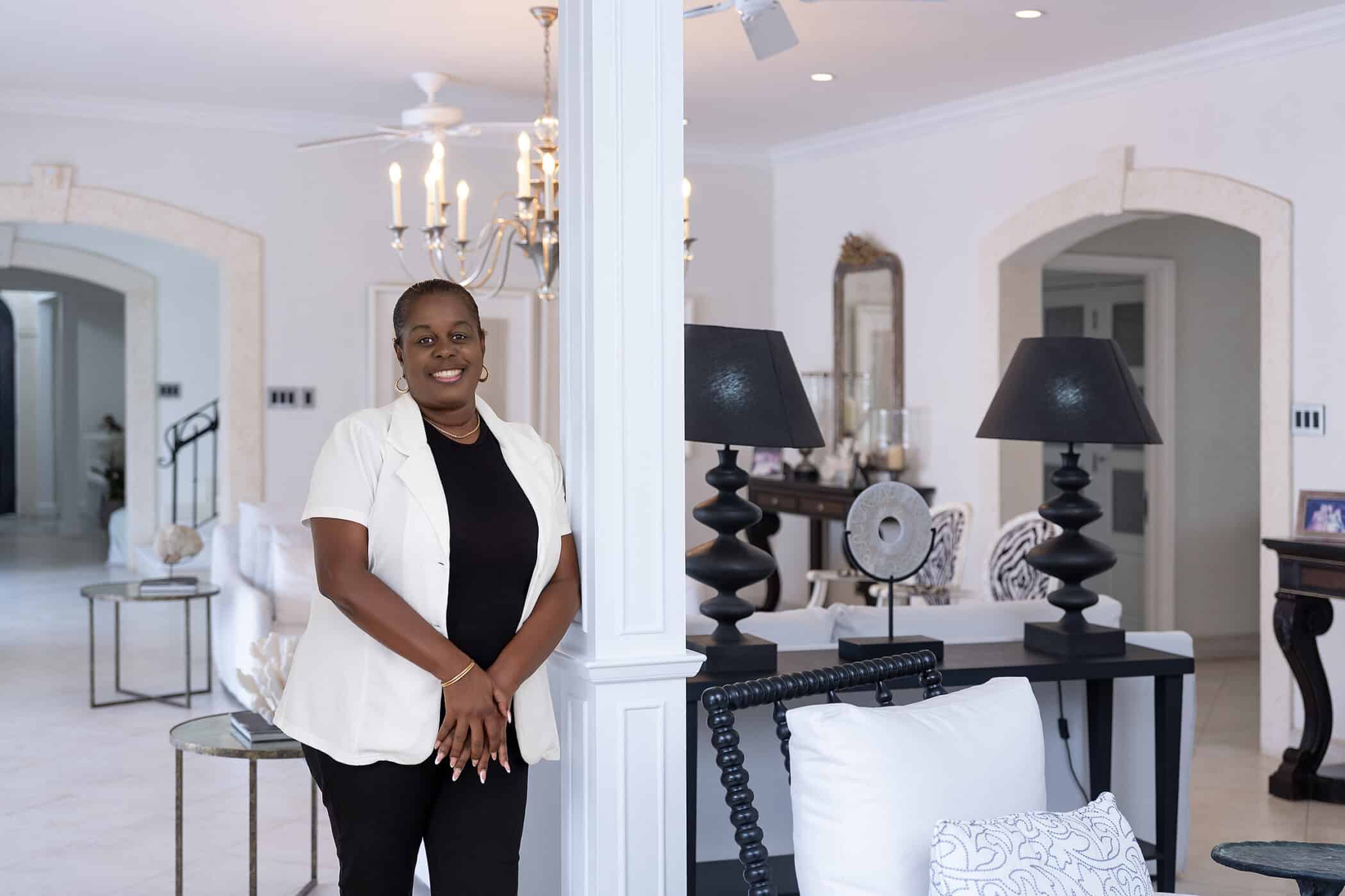 A woman stands smiling in a bright, elegant living room with white walls and modern black and white decor, including lamps, chairs, and a chandelier—her space perfectly capturing the breezy sophistication of the Tradewinds style.