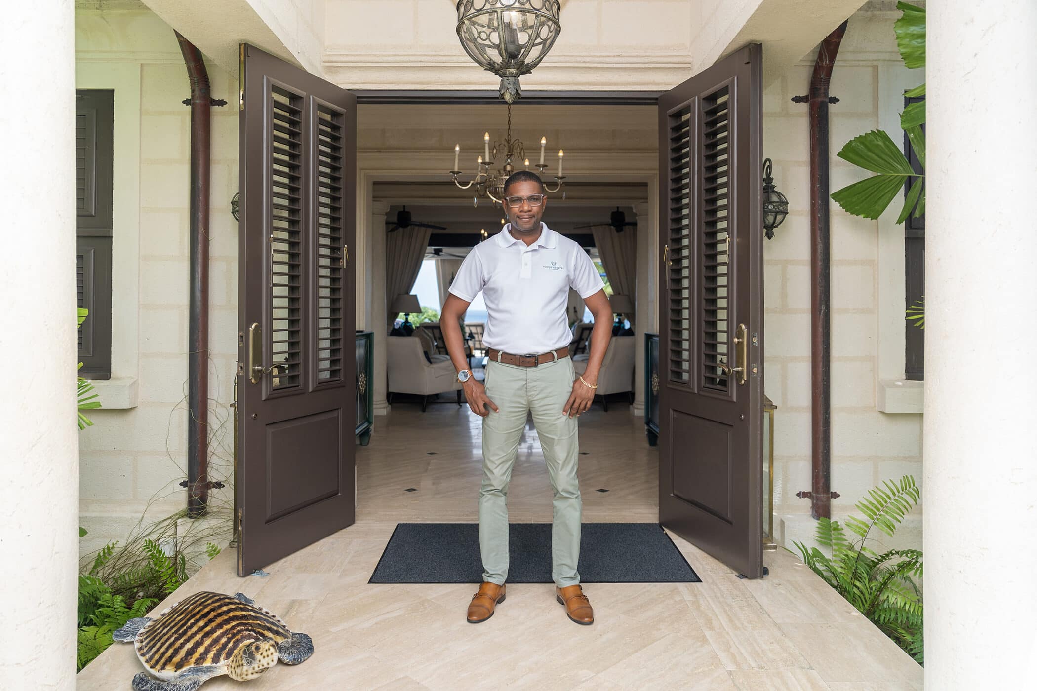 A man wearing glasses, a light gray polo shirt, khaki pants, and brown shoes stands confidently in a doorway of an elegant building called Seaclusion, with ferns and a tortoise sculpture on the floor beside him.