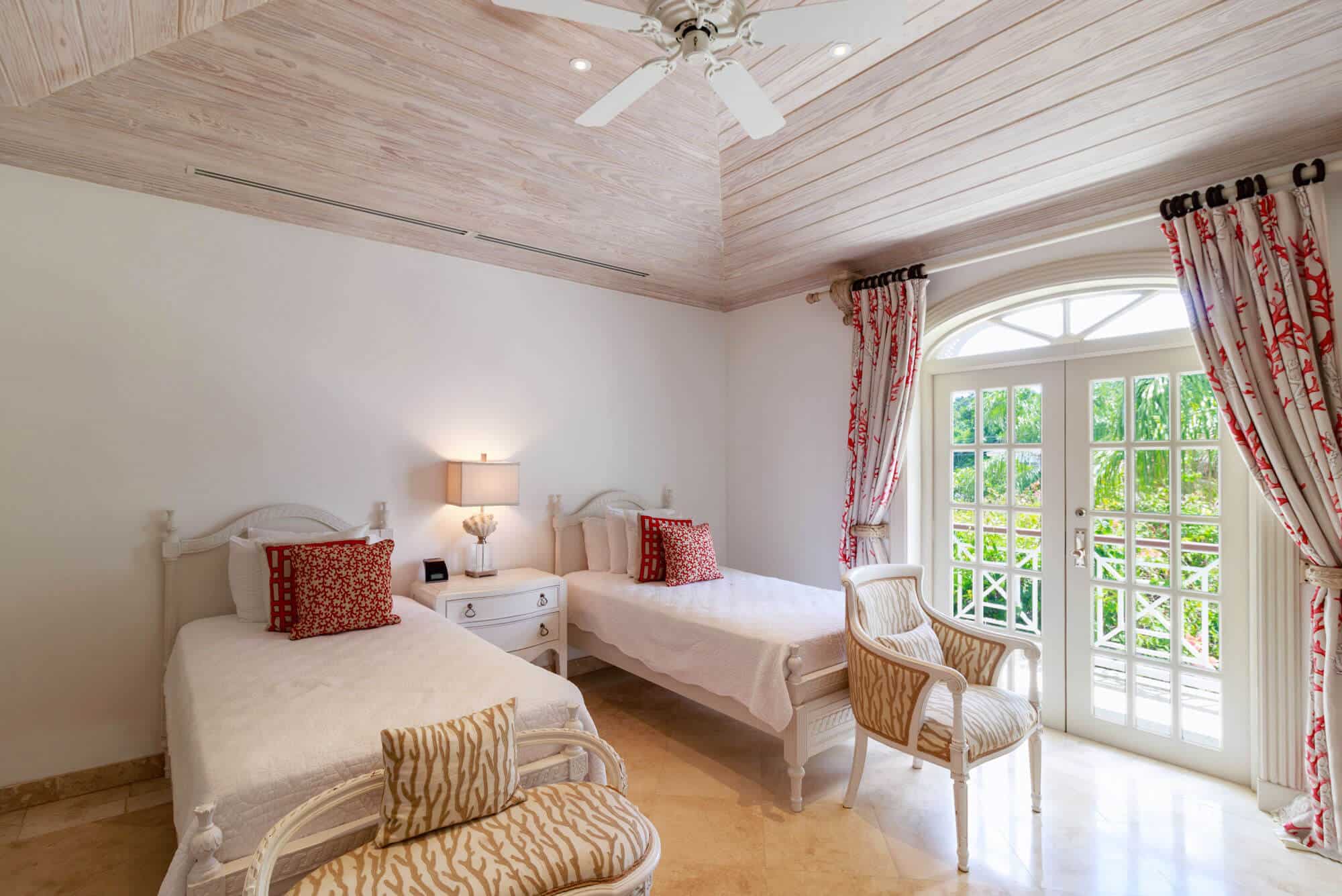 A bright bedroom at Firefly at Old Trees features two single beds, white furniture, patterned cushions, a lamp on a bedside table, an armchair, and French doors with red curtains opening to a view of greenery outside.
