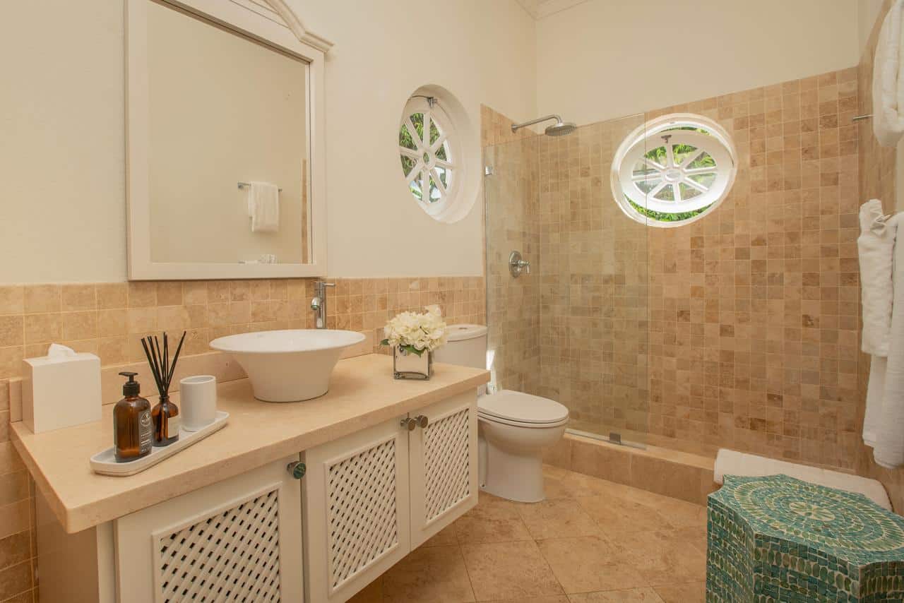 Modern bathroom with a glass shower, beige tiles, and a round window letting in natural light reminiscent of the Aquilae style. A white sink sits on a light countertop, complemented by decorative accessories including bottles and flowers. Towels hang neatly on the wall.