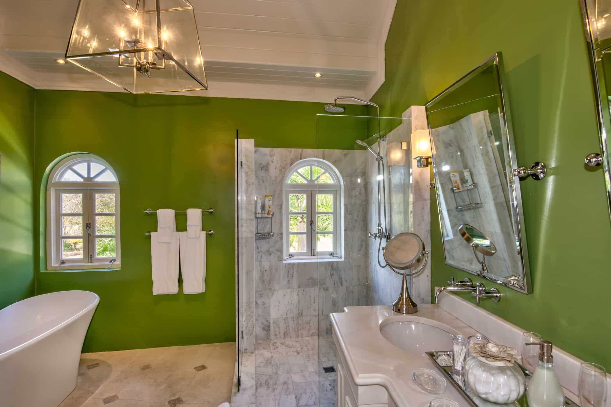A modern bathroom at Casablanca at Sandy Lane features bright green walls, a white freestanding tub, marble shower with glass doors, dual sinks, round mirrors, wall lamps, and two arched windows that let in natural light.