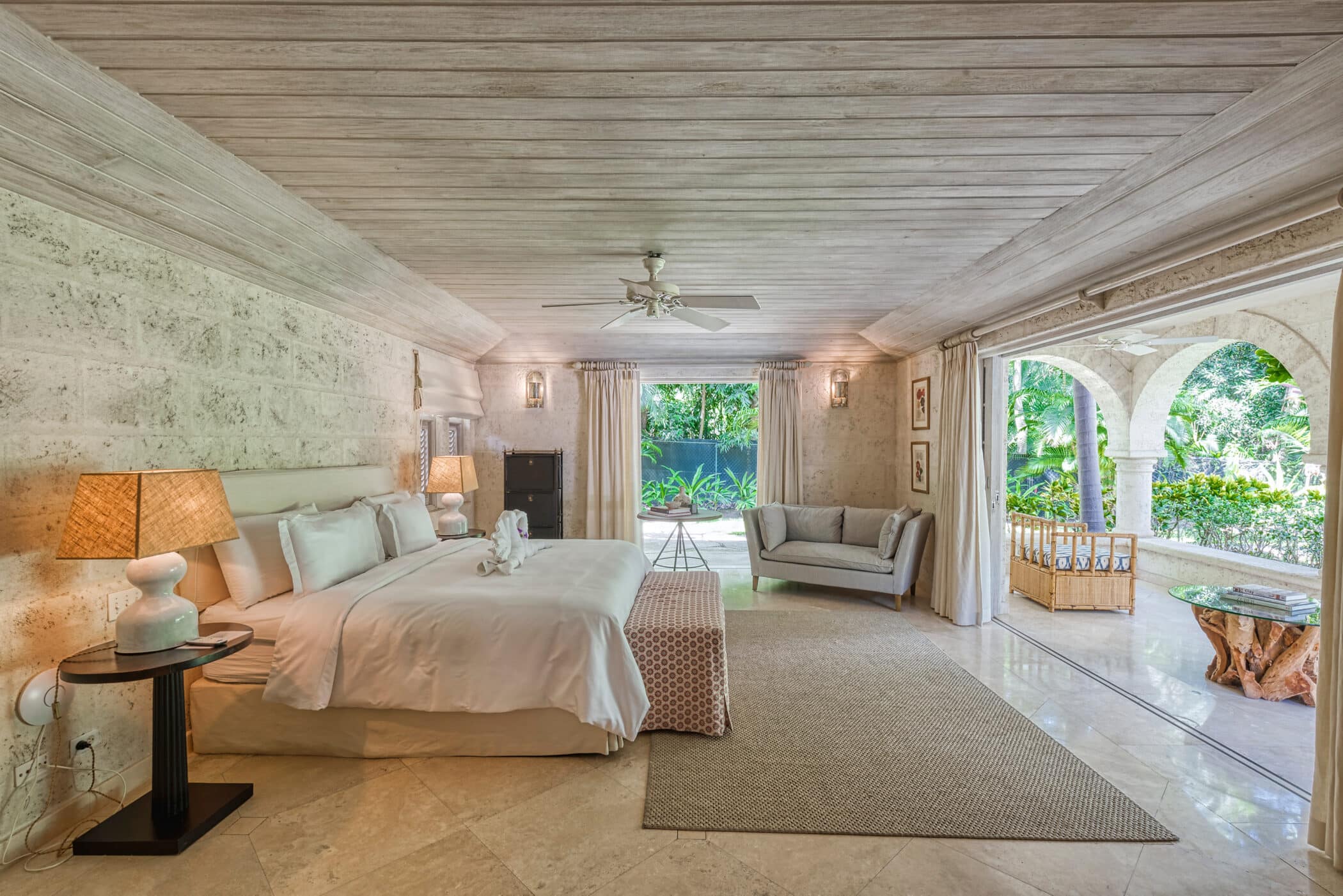 Spacious bedroom at Bluff House and Cottage with a large bed, elegant lamps, sofa, and natural stone walls. The room opens to a lush garden through arched doorways, creating a serene and airy atmosphere.