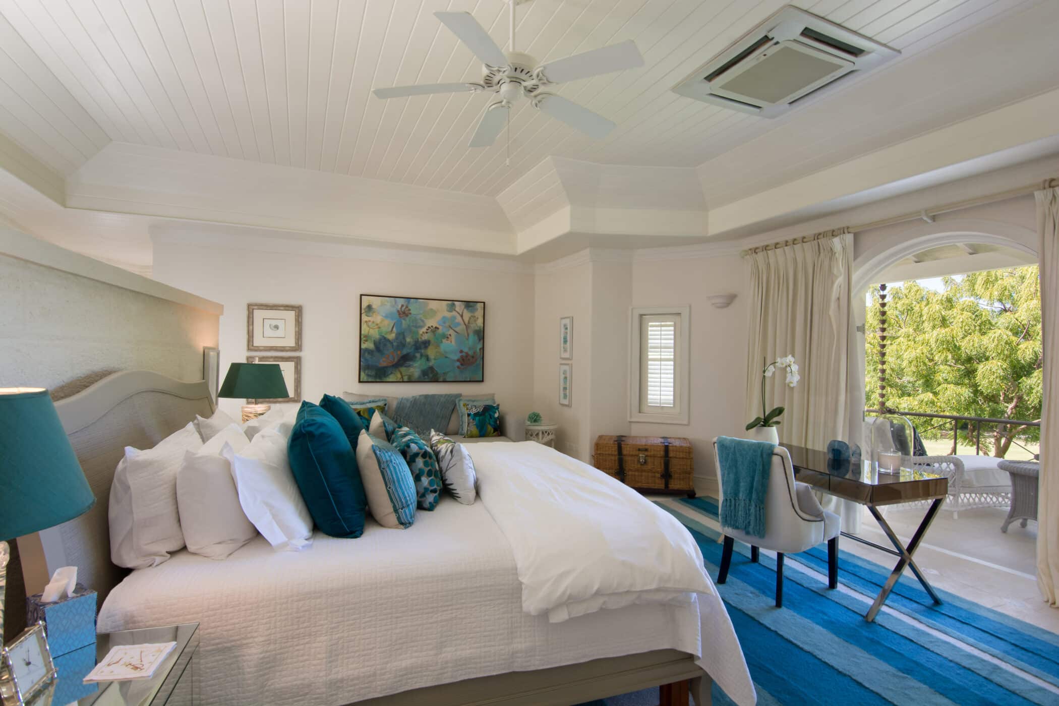 Bright bedroom at Casablanca at Sandy Lane with a large bed, many blue accent pillows, a blue rug, artworks, a desk near a window, and glass doors leading to a green outdoor area. Ceiling fan and natural light enhance the spacious feel.