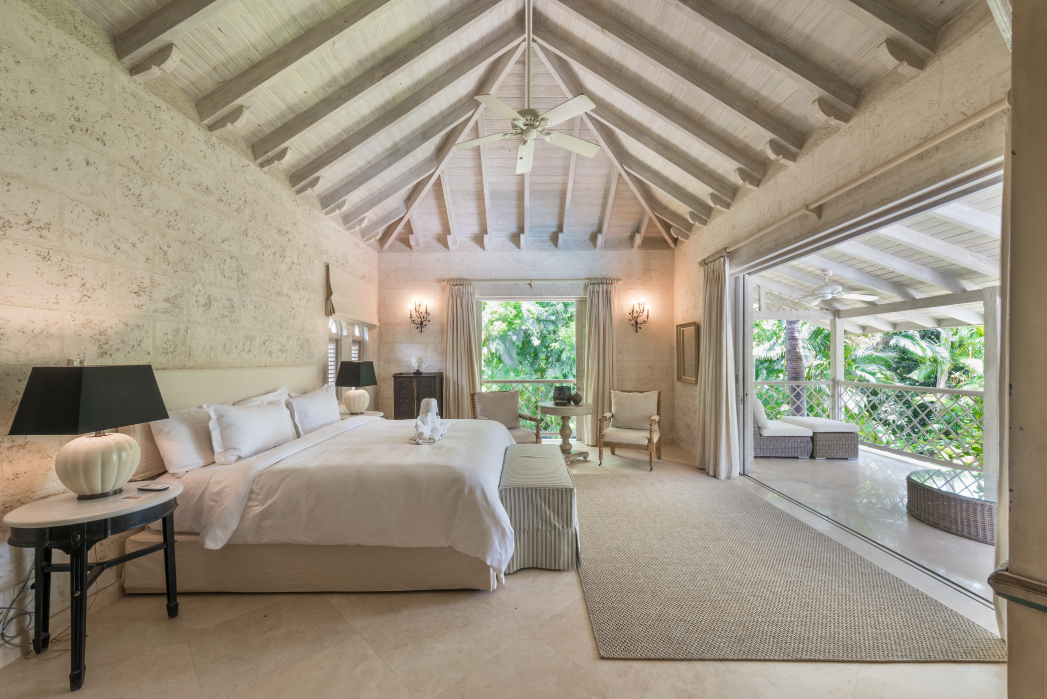 Spacious, airy bedroom at Bluff House and Cottage with a vaulted ceiling, large bed, side tables, and lamps. Sliding glass doors open to a covered balcony with seating, overlooking lush green trees. Natural light fills the room.
