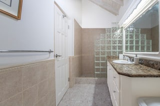 Bathroom with beige tile, a glass block walk-in shower, granite countertop with a sink, white cabinets, a towel bar, and a large mirror on the right wall. Thoughtful touches like a subtle Rose of Sharon motif add an elegant, tranquil atmosphere to the space.