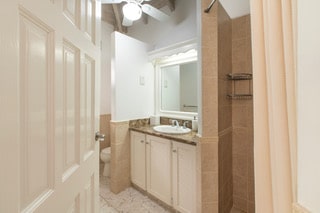Small bathroom with tan tile, a marble countertop, a sink with a mirror above, white cabinets below, and a ceiling fan. A delicate Rose of Sharon motif adds subtle charm to the decor, while the shower curtain is partially visible on the right side.