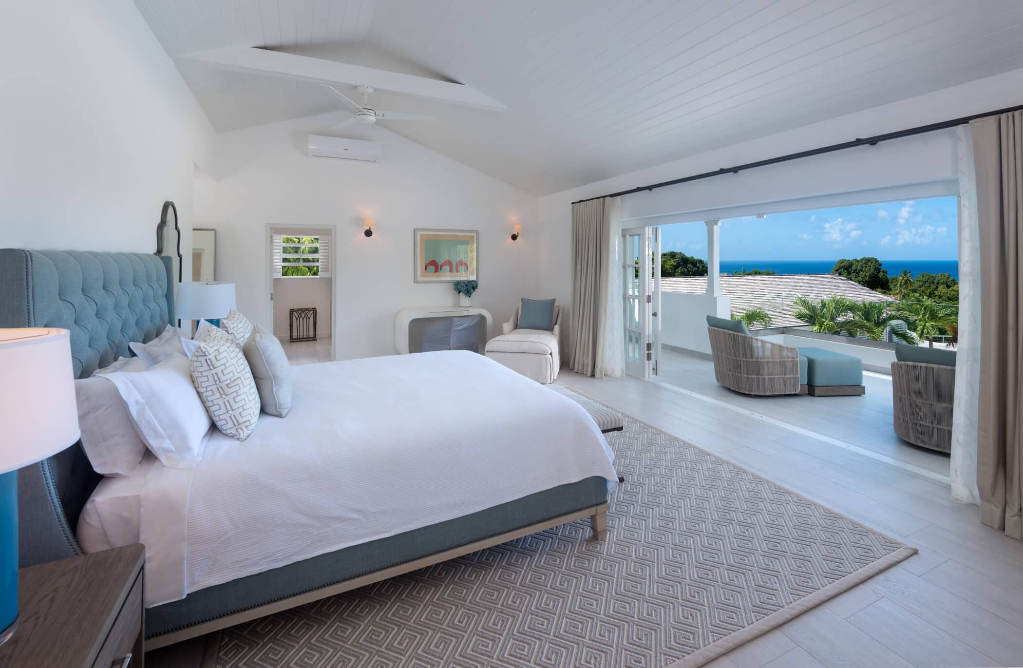Spacious, modern bedroom with a large bed, armchair, and outdoor patio offering ocean views through open glass doors. Decor is light and airy with neutral tones, creating a tranquil retreat where you can unwind and enjoy the refreshing sea breeze.