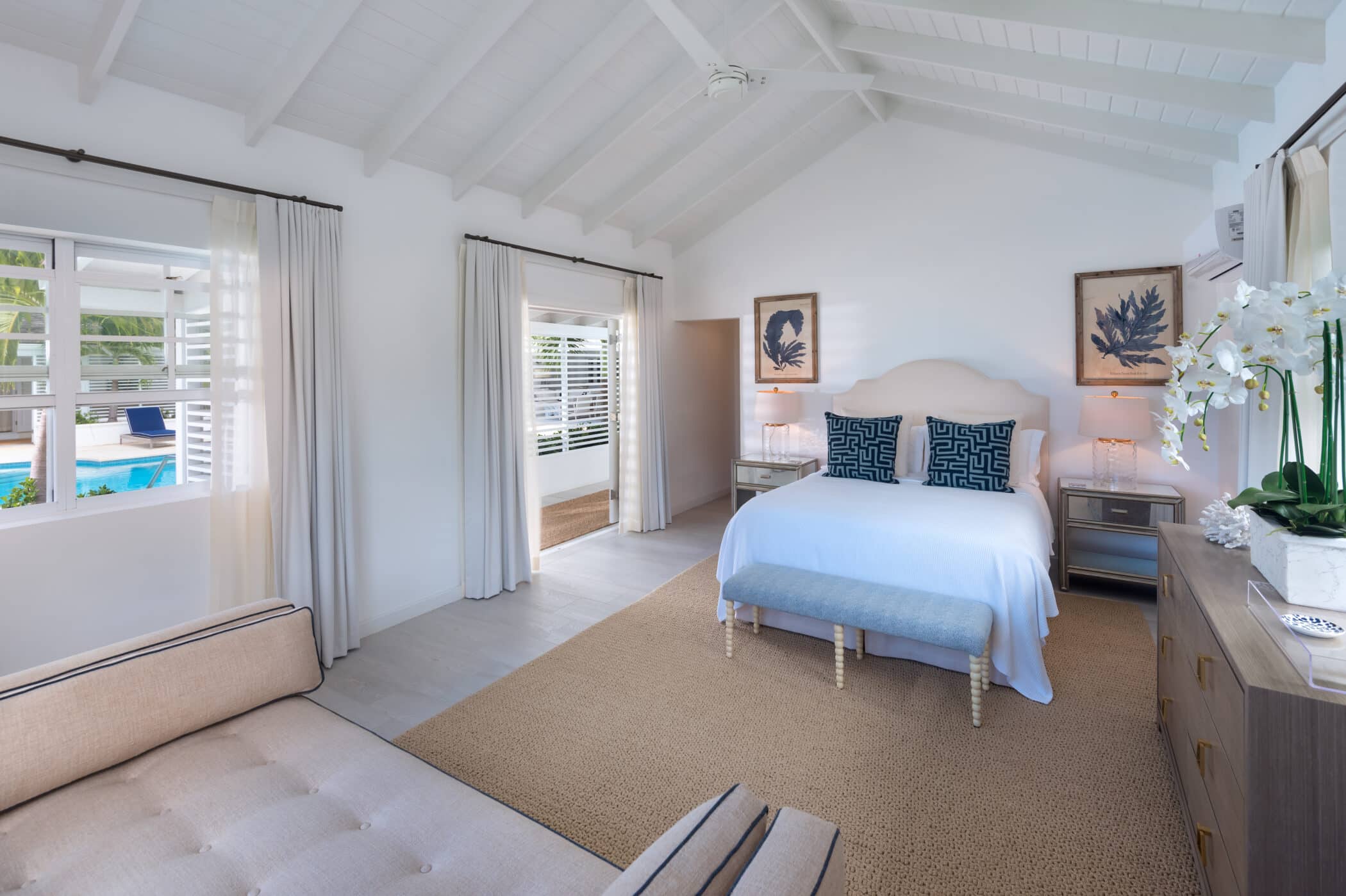 Bright bedroom with white walls, a cozy bed with blue Sea Breeze accents, large windows with curtains, a beige sofa, a dresser with flowers, and a view of a pool outside. Artwork and lamps decorate the space.