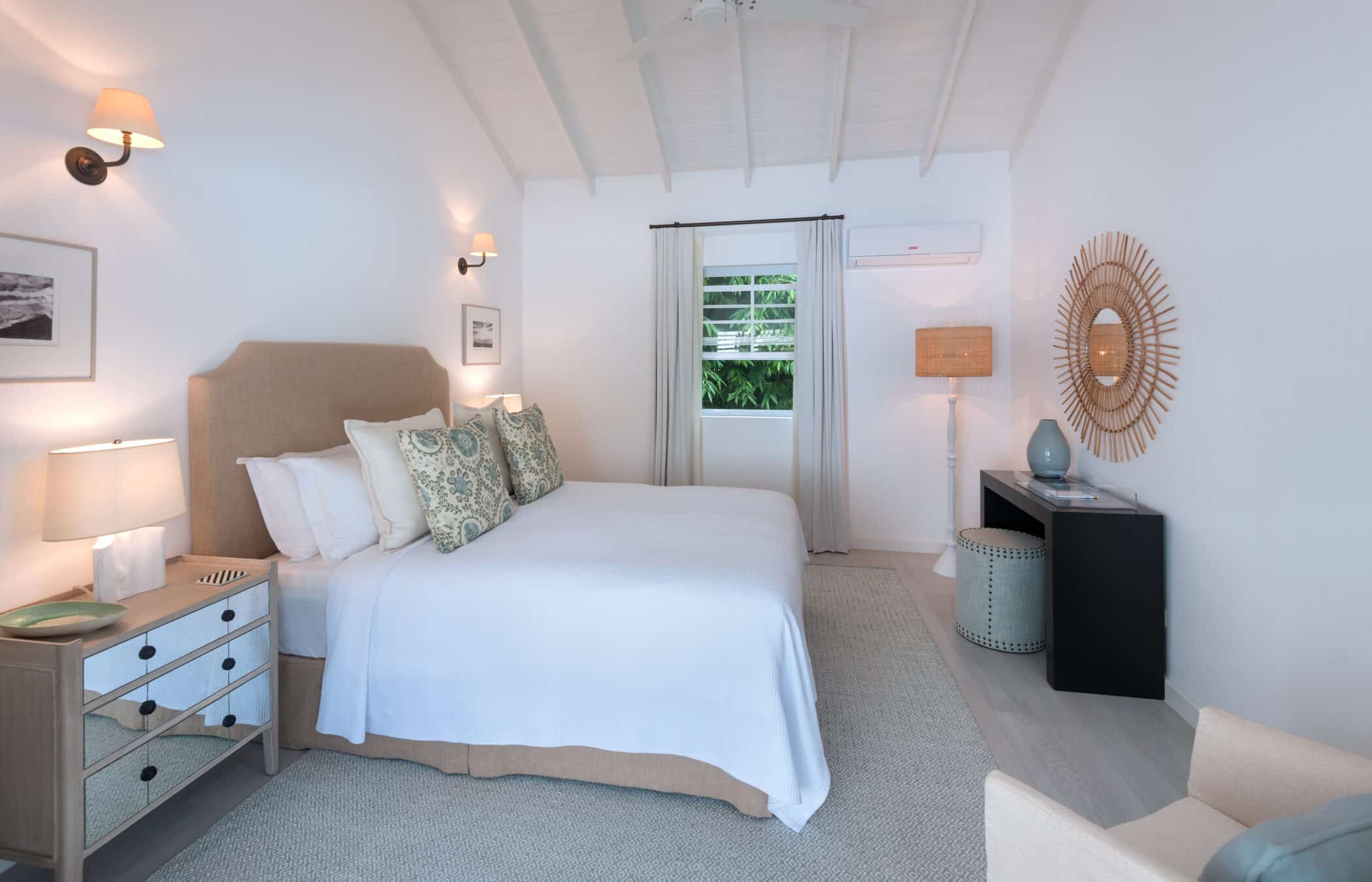 Bright bedroom with a beige bed, patterned pillows, two nightstands, lamps, a decorative mirror, a black console, and a window with sheer curtains showing greenery outside. White walls and ceiling create an airy feel reminiscent of a gentle Sea Breeze.