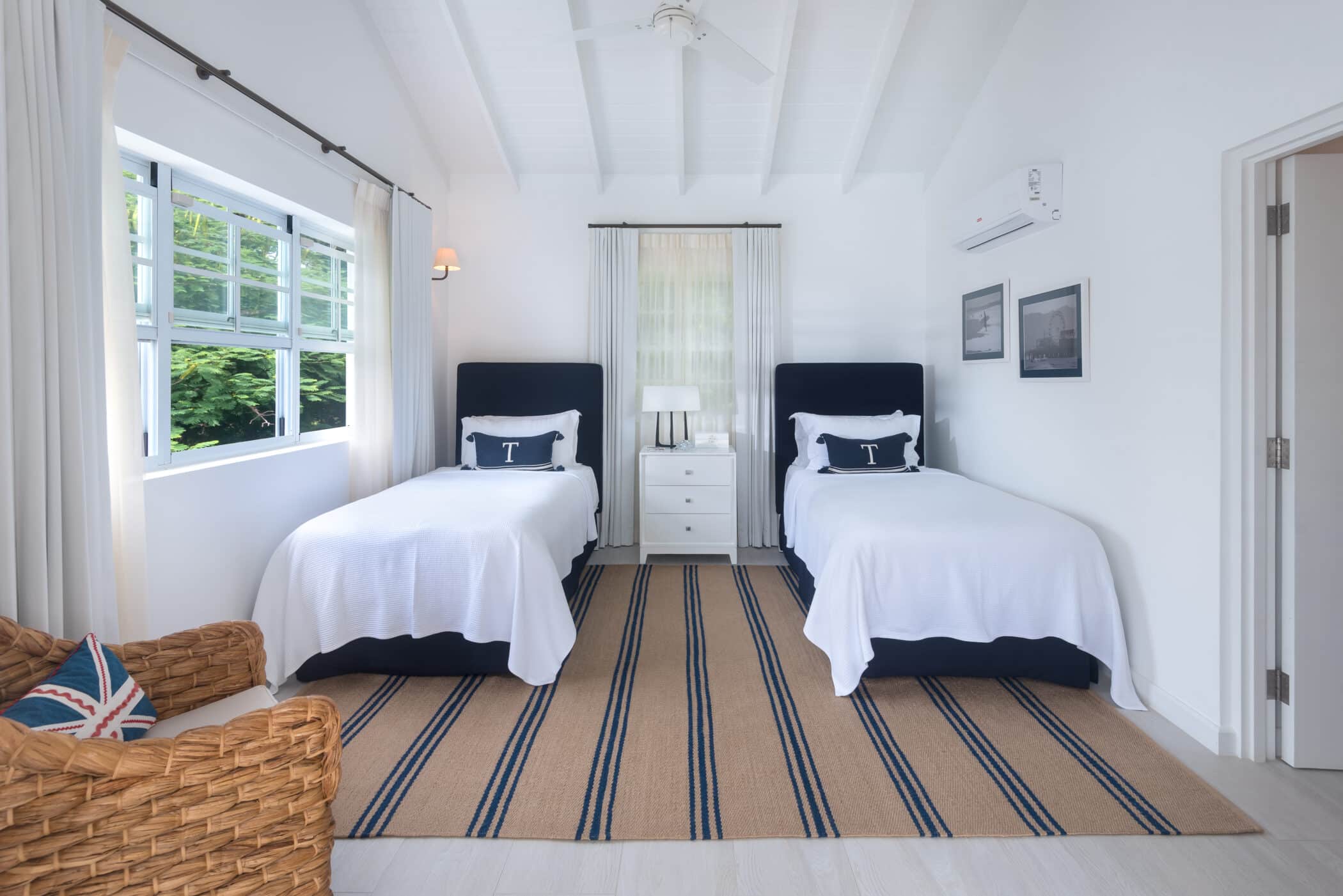 Bright bedroom with two twin beds, white bedding, a striped rug, wicker chair, and large window with curtains inviting in a gentle sea breeze. White walls and ceiling create an airy atmosphere, complemented by a nightstand and lamp between the beds.