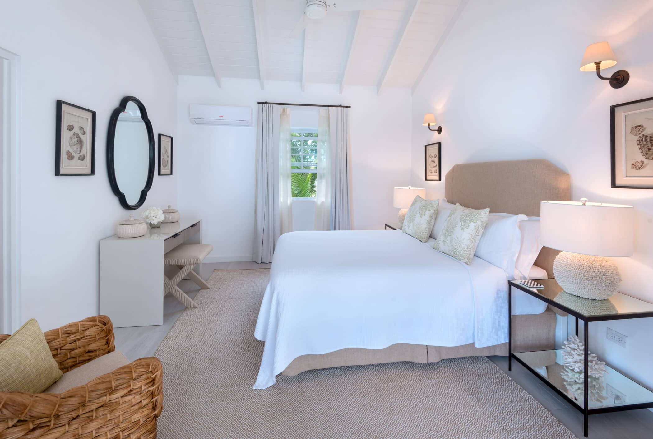 Bright bedroom with a beige bed, white bedding, wicker chair, wall art, bedside lamps, desk with mirror, two vases, and a window with light curtains letting in natural light—a tranquil retreat inspired by the refreshing feel of a Sea Breeze.
