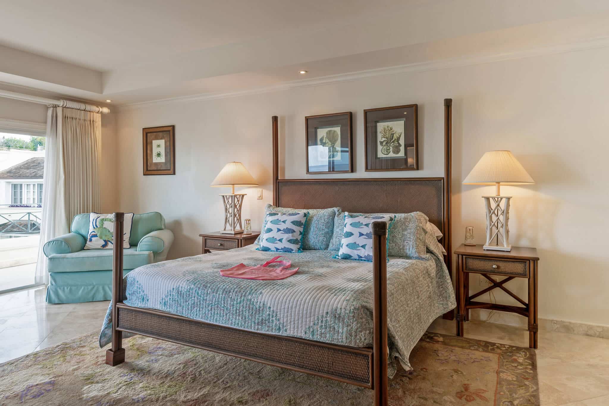 A bright bedroom at The Penthouse at Schooner Bay features a large bed with patterned pillows and quilt, two bedside tables with lamps, framed art above the bed, and a blue armchair near a window showing a balcony outside.