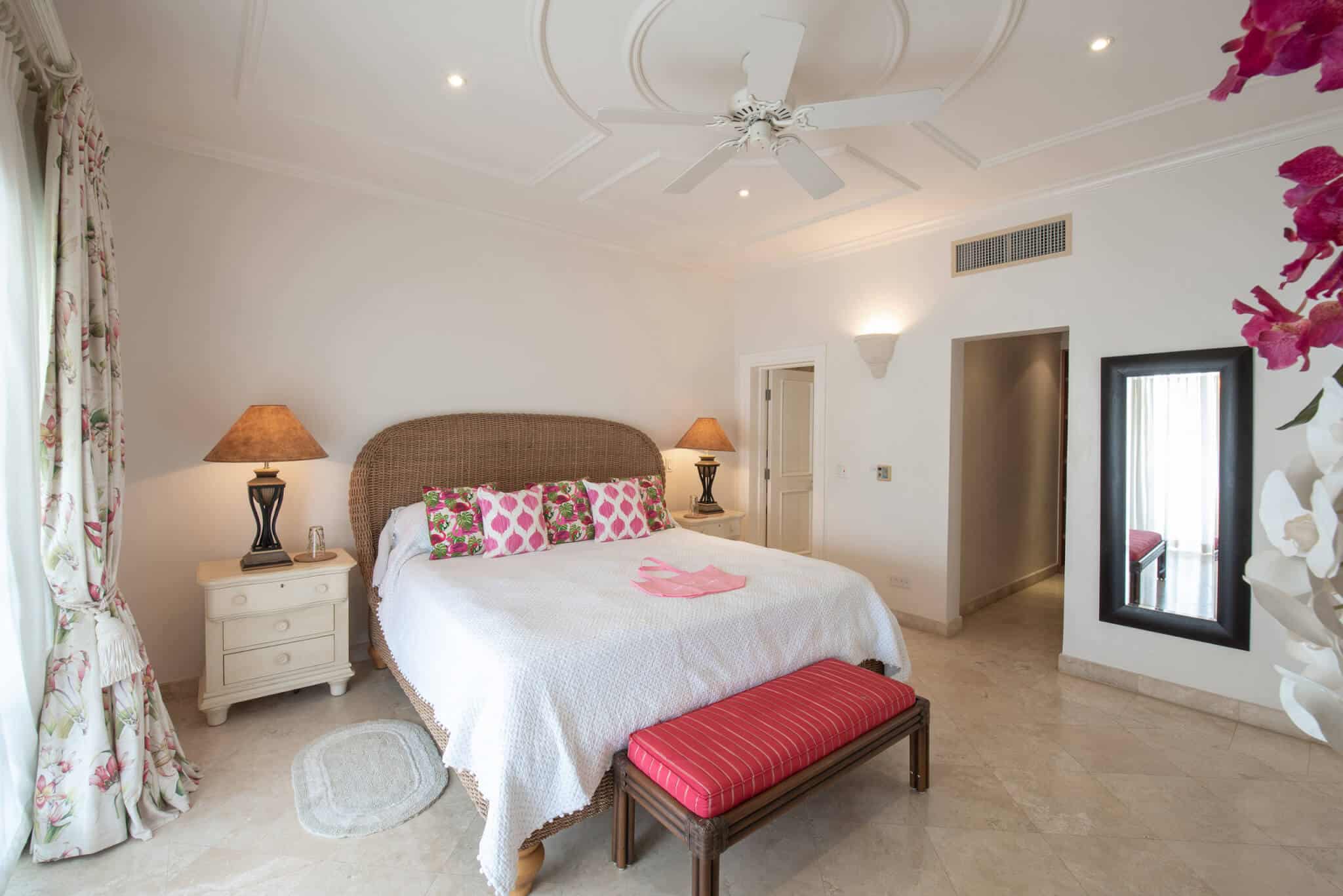 A bright bedroom in The Penthouse at Schooner Bay features a large bed, patterned pillows, two lamps, a red bench, a mirror, and floral curtains. Light walls, tile floor, and a ceiling fan create an airy, inviting atmosphere.