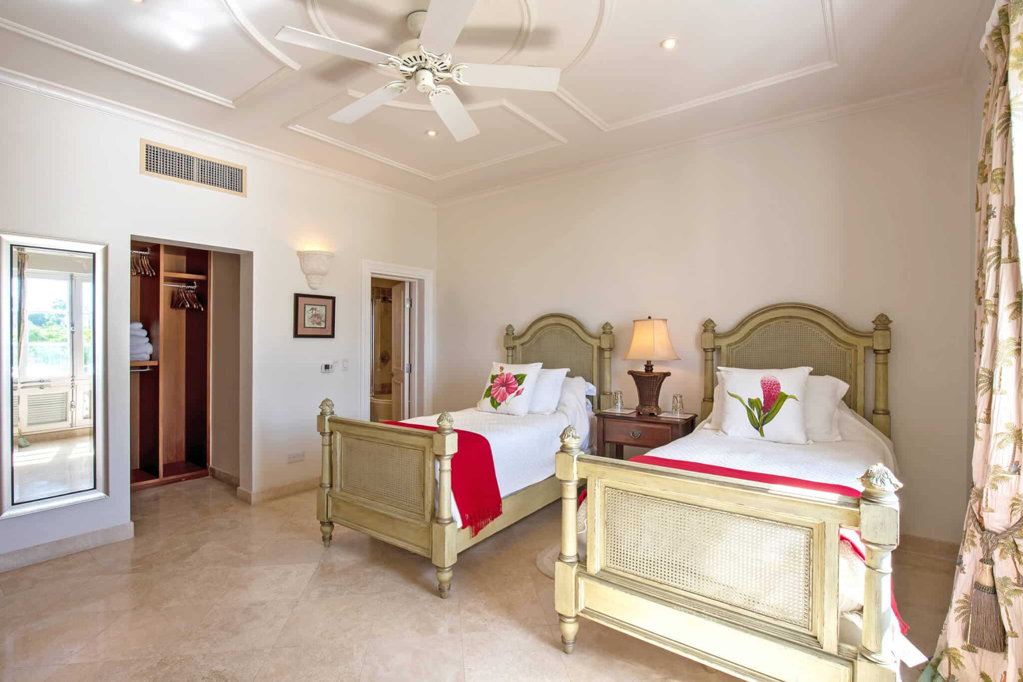 Bright bedroom with two twin beds, floral decorations, a bedside lamp, ceiling fan, and an open closet. Large window with floral curtains lets in natural light. Light-colored walls and tile flooring complete the room—perfectly complementing the elegant ambiance of The Penthouse at Schooner Bay.