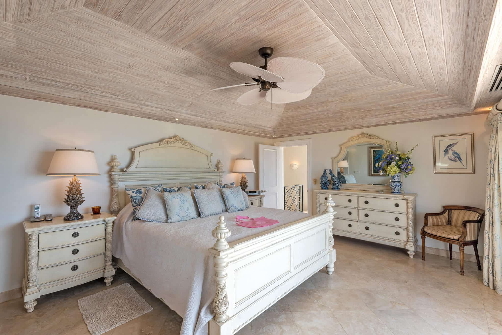 Elegant bedroom at The Lookout at Schooner Bay with a white wooden bed, matching nightstands and dresser, cozy armchair, lamps, wall art, and a ceiling fan, all bathed in natural light from a window out of frame.