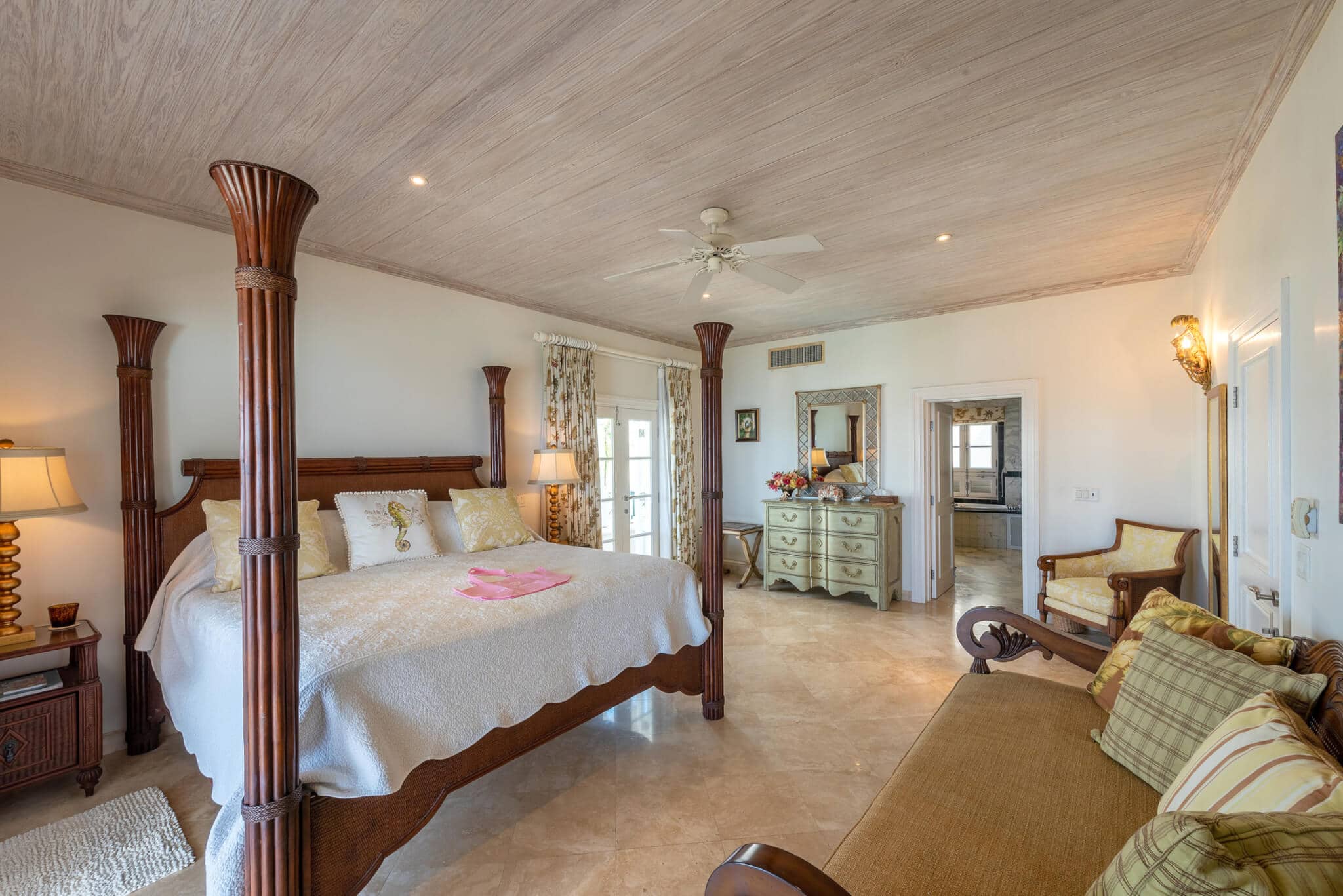 Spacious bedroom at The Lookout at Schooner Bay featuring a four-poster bed, elegant wooden furniture, cushioned bench, marble floor, and large windows with curtains letting in natural light.