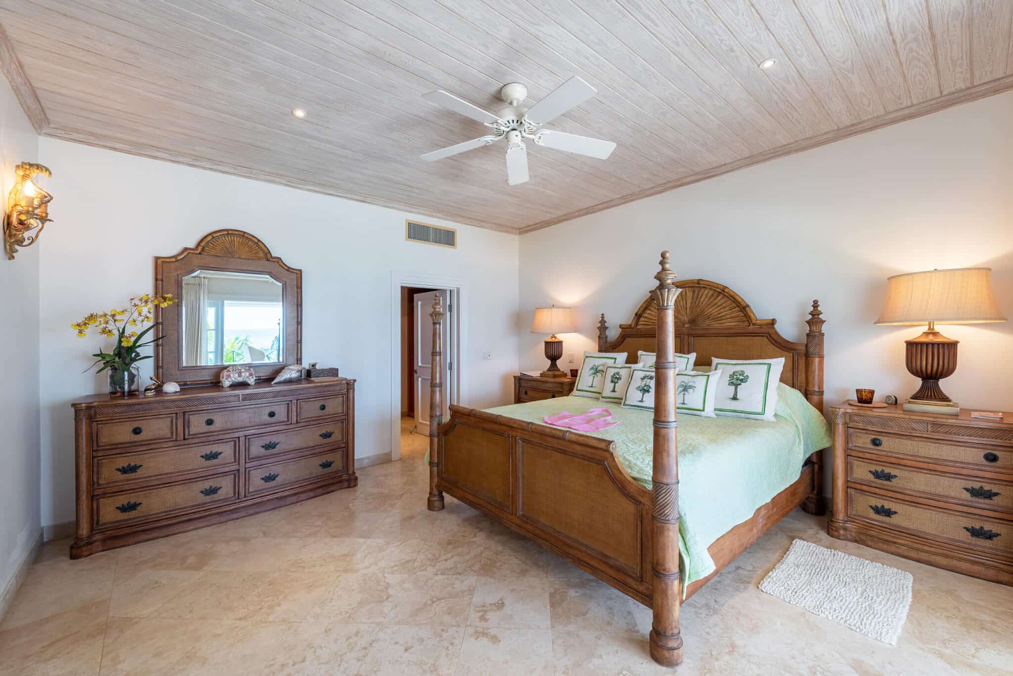 Spacious bedroom at The Lookout at Schooner Bay featuring a wooden bed, matching dresser, two lamps, floral decor, a ceiling fan, light tile floor, and a window with an outside view.