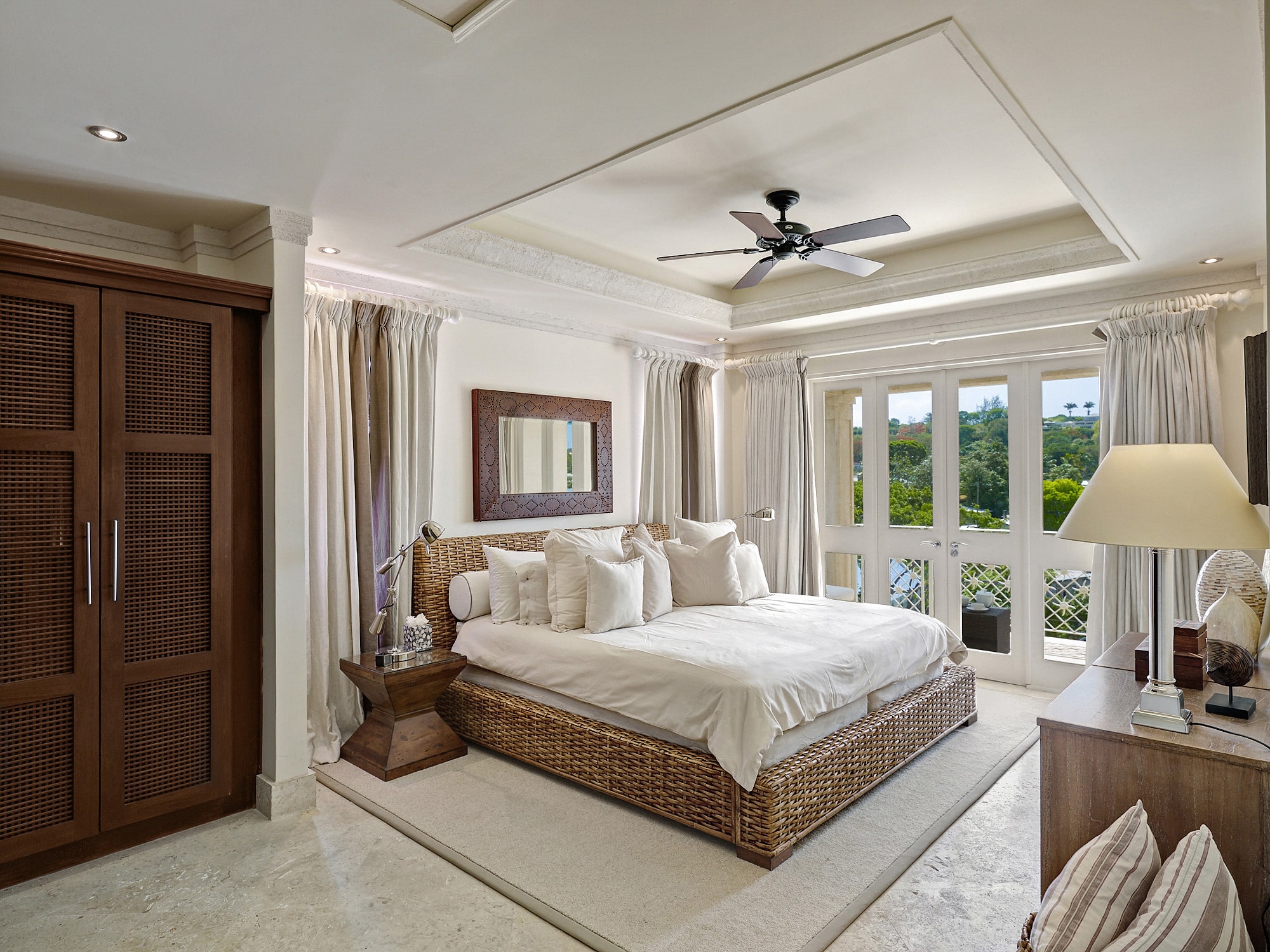 Bright bedroom in the Smugglers Cove Penthouse, featuring a wicker bed with crisp white bedding, large windows with curtains, a ceiling fan, wooden wardrobe, bedside tables, and a dresser with a lamp. A mirror hangs above the bed while lush greenery is visible just outside.