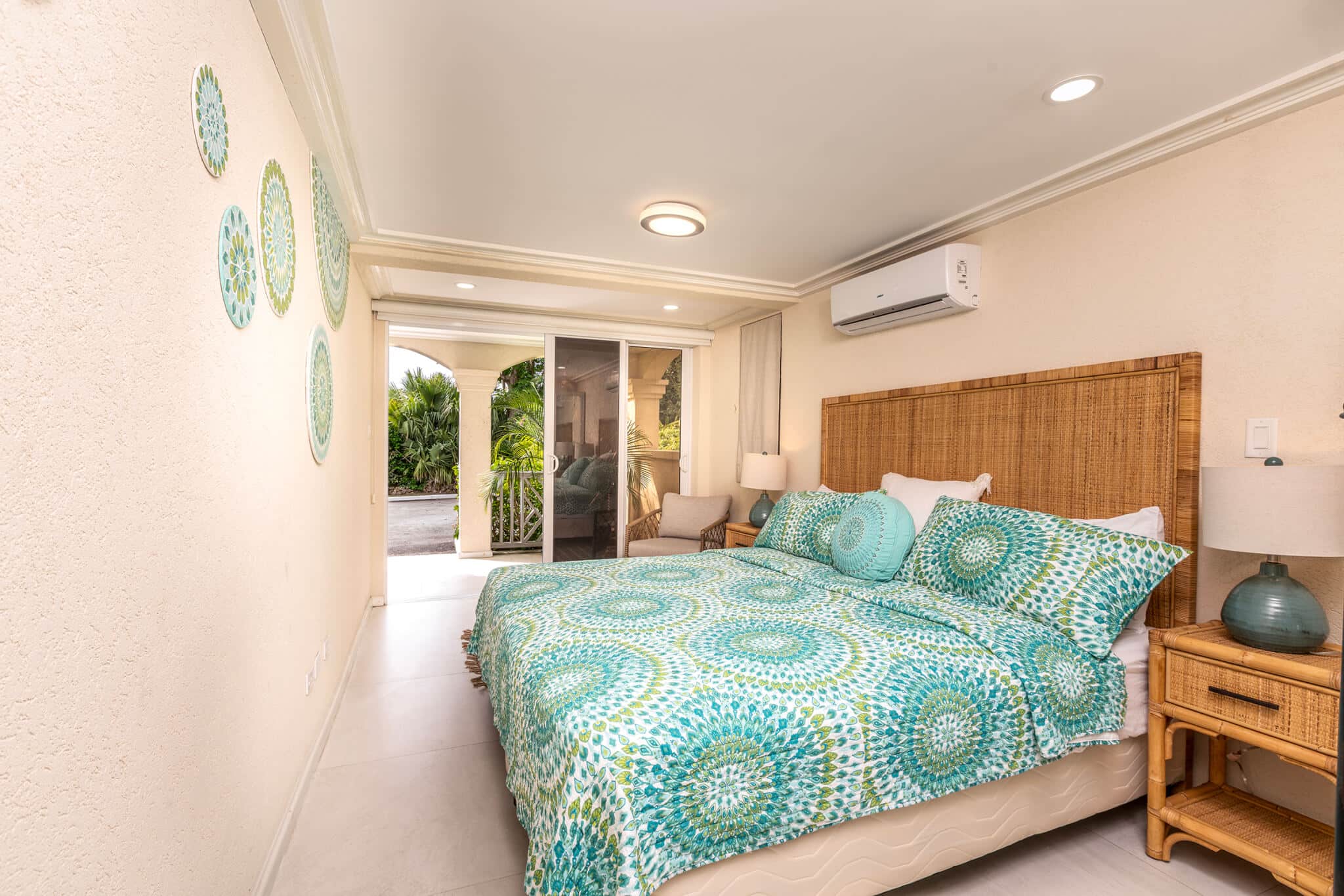 Bright Casabella bedroom with a large bed, patterned green and blue bedding, wicker headboard, nightstand with lamp, wall art, and sliding glass doors opening to a patio with lush greenery outside.
