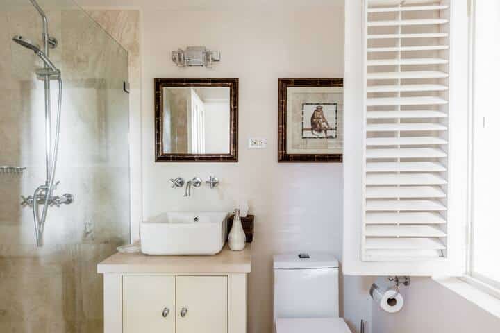 Bright, modern bathroom at Casablanca at Sandy Lane with a glass shower, white sink on a cabinet, wall-mounted faucet, mirror, framed bird art, white toilet, toilet paper roll, and sunlight streaming through white shutters.