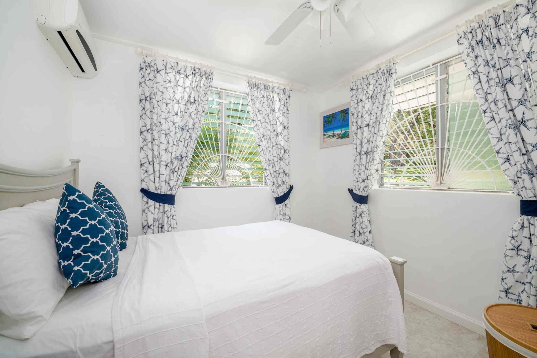 Bright bedroom with a white bed, blue-patterned pillows, floral curtains, a ceiling fan, wall art by Halle Rose, and barred windows letting in natural light. An air conditioner is above the bed and a small round wooden table is beside it.