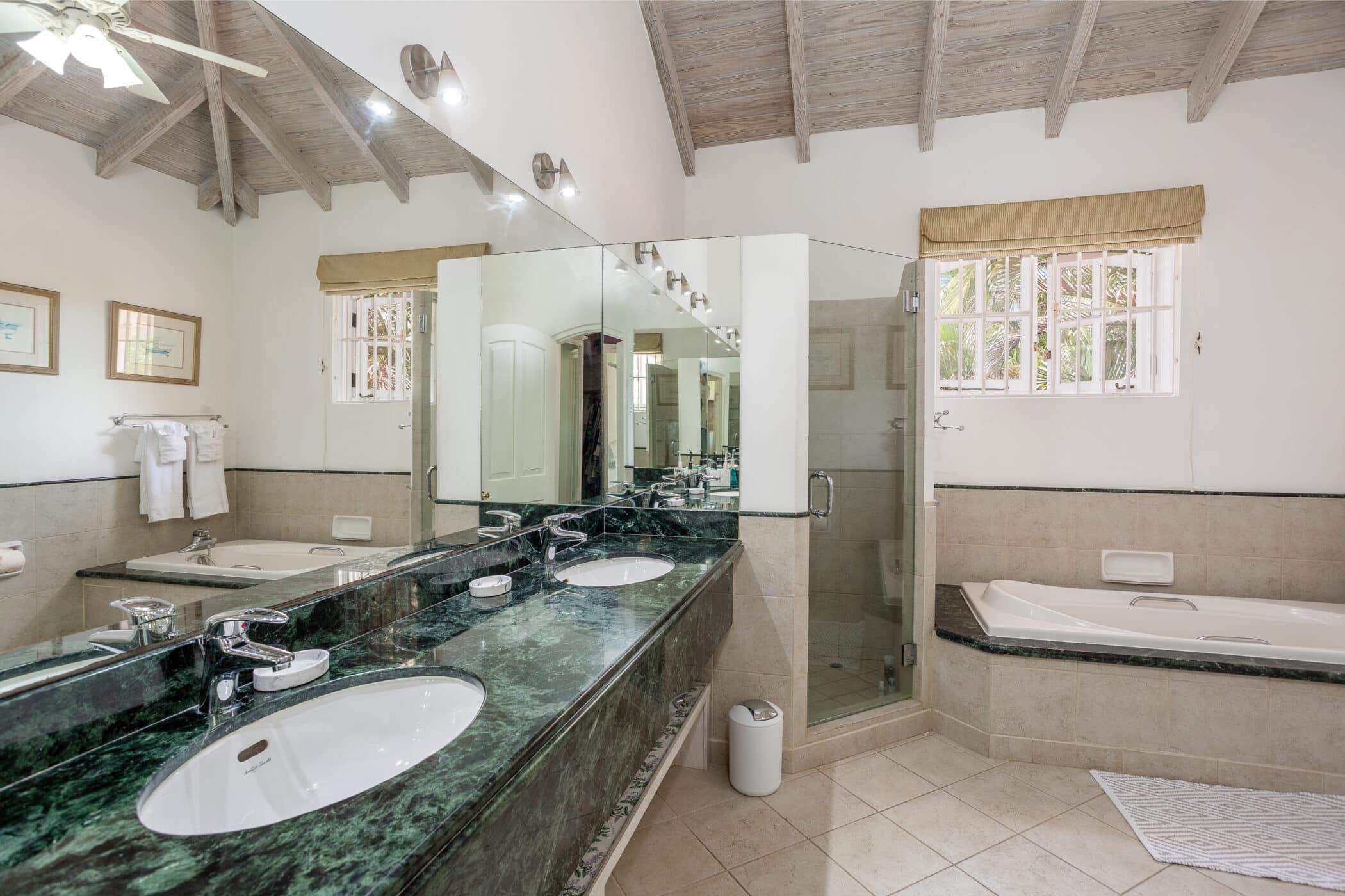 Spacious bathroom with a double sink vanity, large mirror, green marble countertop, glass-enclosed shower, bathtub with tile surround, tiled floor, and beamed ceiling with windows letting in natural light.