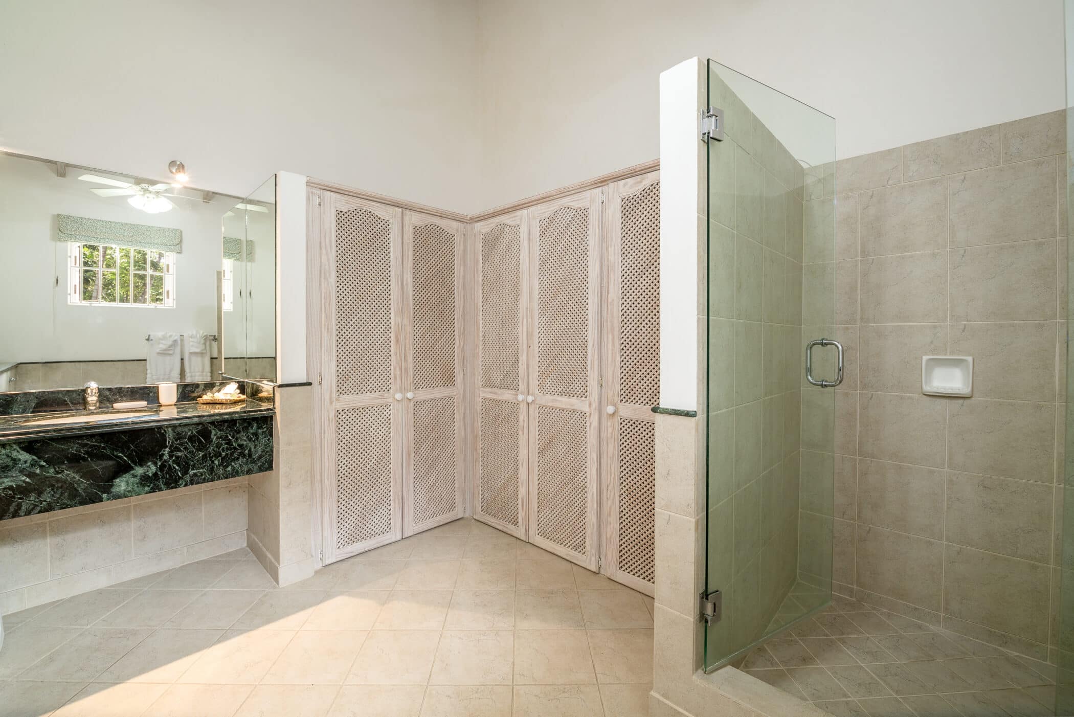 A spacious, modern bathroom with beige tile flooring, a glass-enclosed shower, a green marble countertop with a sink, large mirrored wall, and white lattice-front cabinets. A window and ceiling fan are visible.