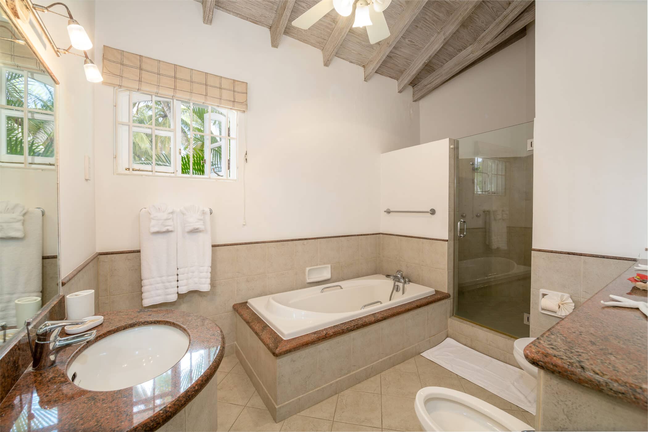 Bright bathroom with a bathtub, glass-enclosed shower, sink with mirror, towels on a rack, and a window with natural light. The room has beige tiles, granite countertops, and a vaulted wooden ceiling.