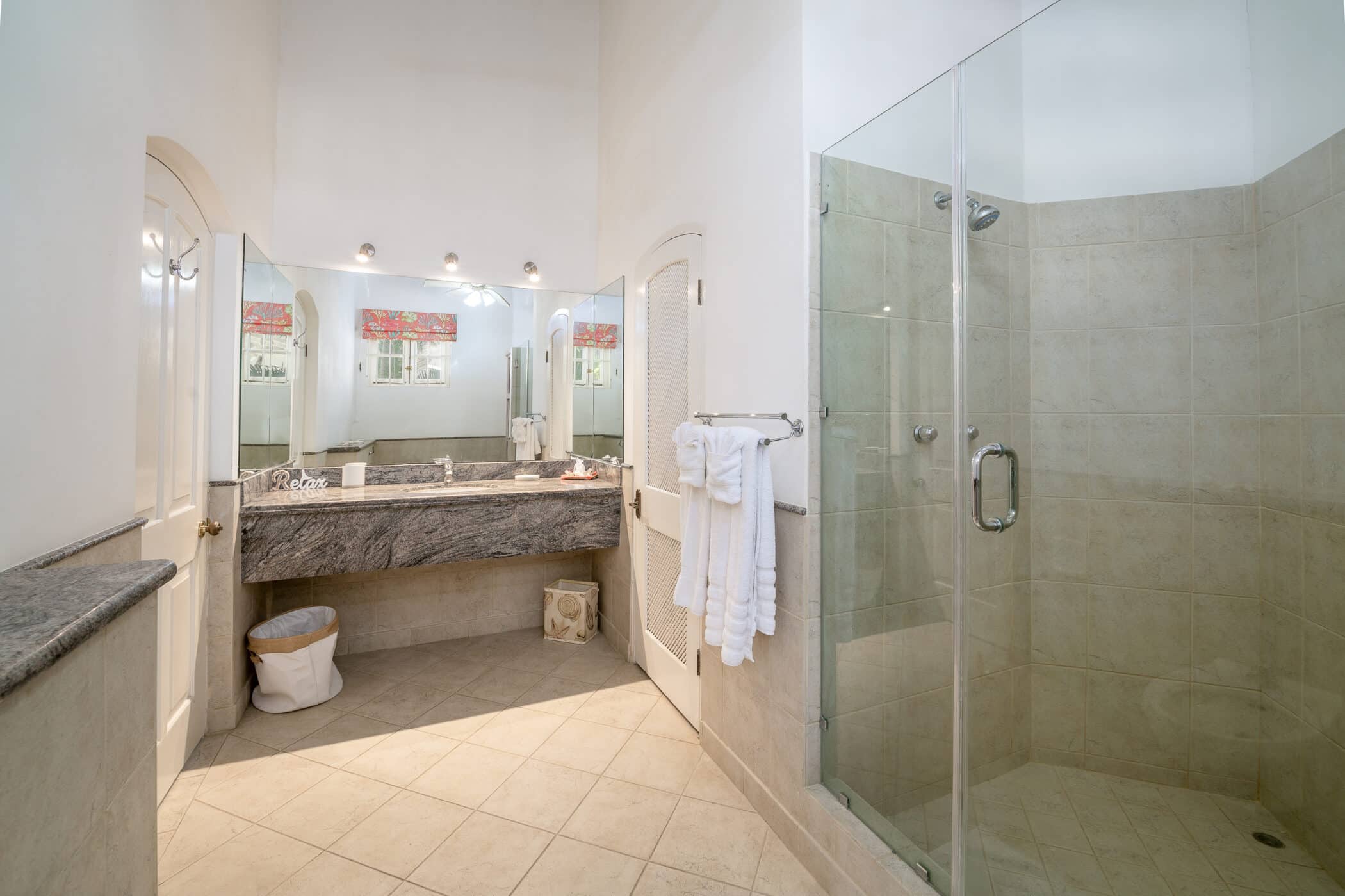 Spacious bathroom with a glass-enclosed shower, marble countertop vanity with towels, large mirrors, tiled floor and walls, and white walls with bright lighting.