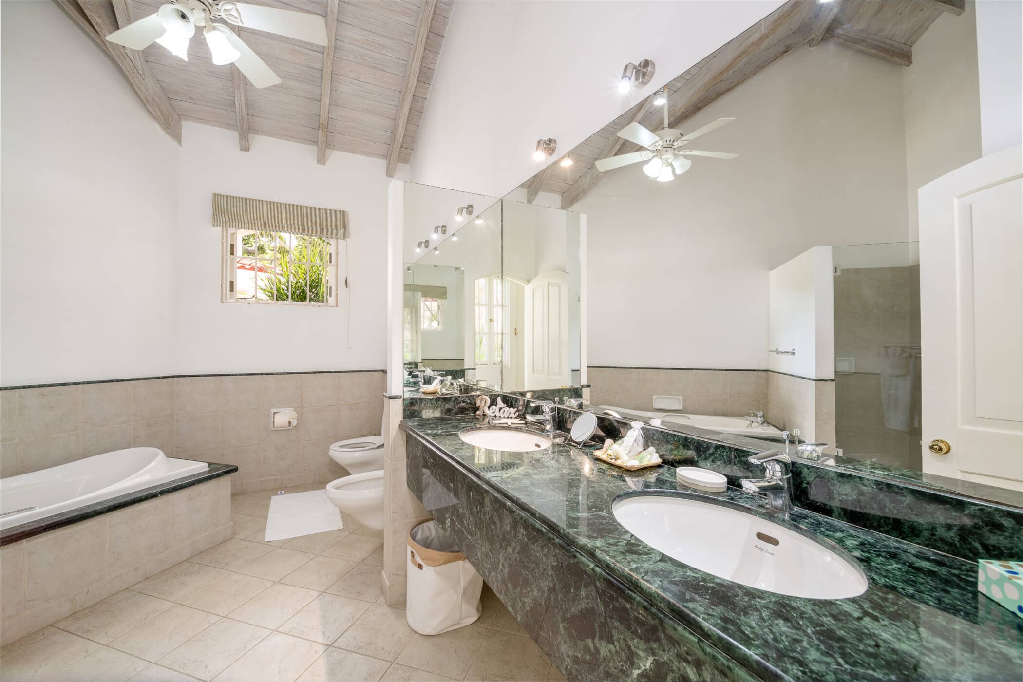 Spacious bathroom with green marble double sink, large mirror, bathtub, toilet, and bidet. High vaulted ceiling with fans, tile flooring, and natural light from a window above the tub.