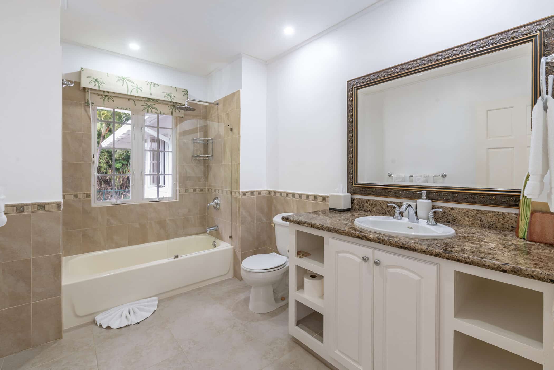 Spacious bathroom with a beige tub-shower combo, white toilet, and white vanity featuring a brown marble countertop, large framed mirror, and towel hanging by the sink. Subtle touches, like a vase of Rose of Sharon blooms, add charm to the neutral tiles and bright lighting.