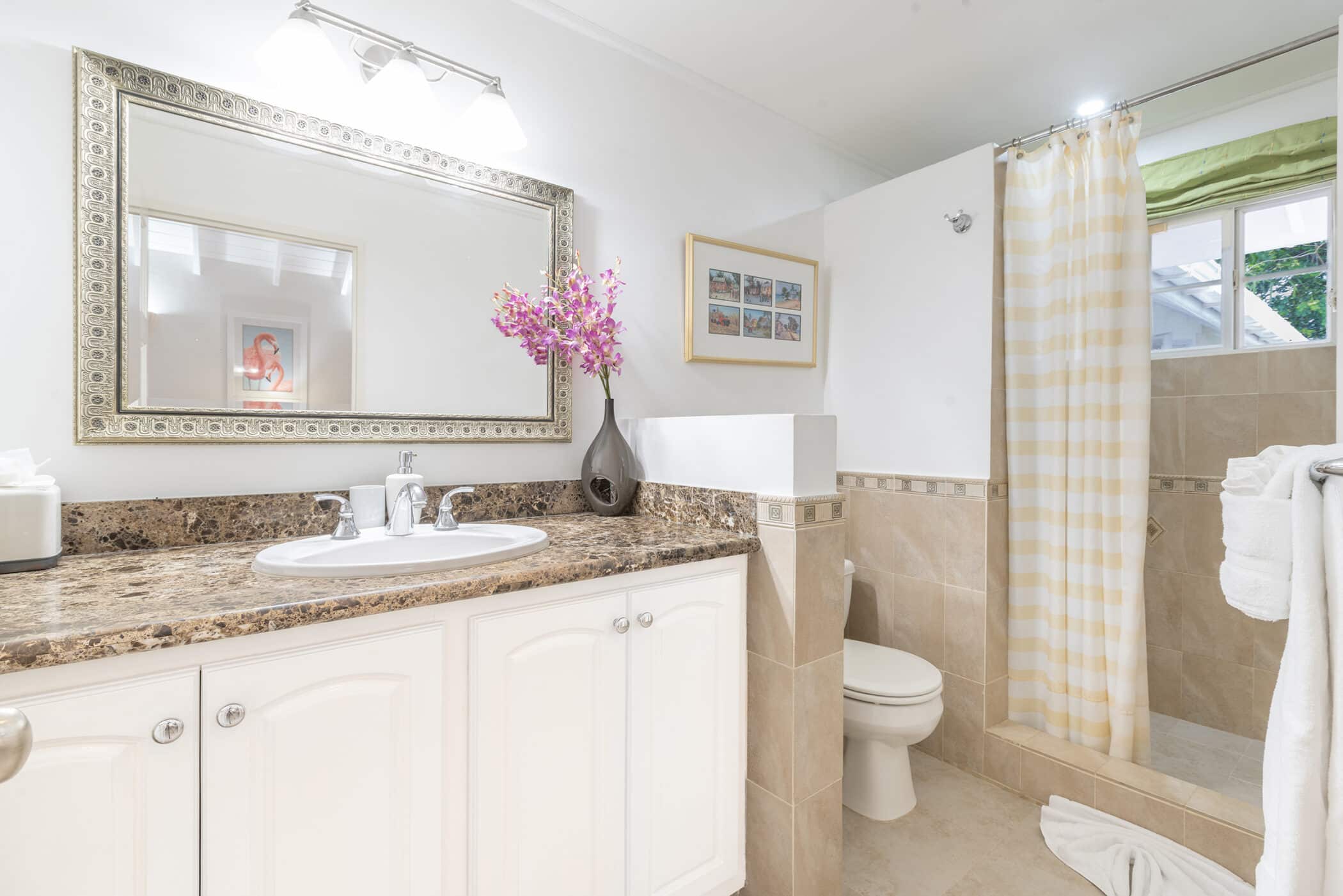 Bright bathroom with a marble counter, sink, and mirror above. Pink Rose of Sharon flowers in a vase add a delicate touch, while a shower with a yellow-striped curtain, toilet, towel rack, and framed art decorate the white and beige room.