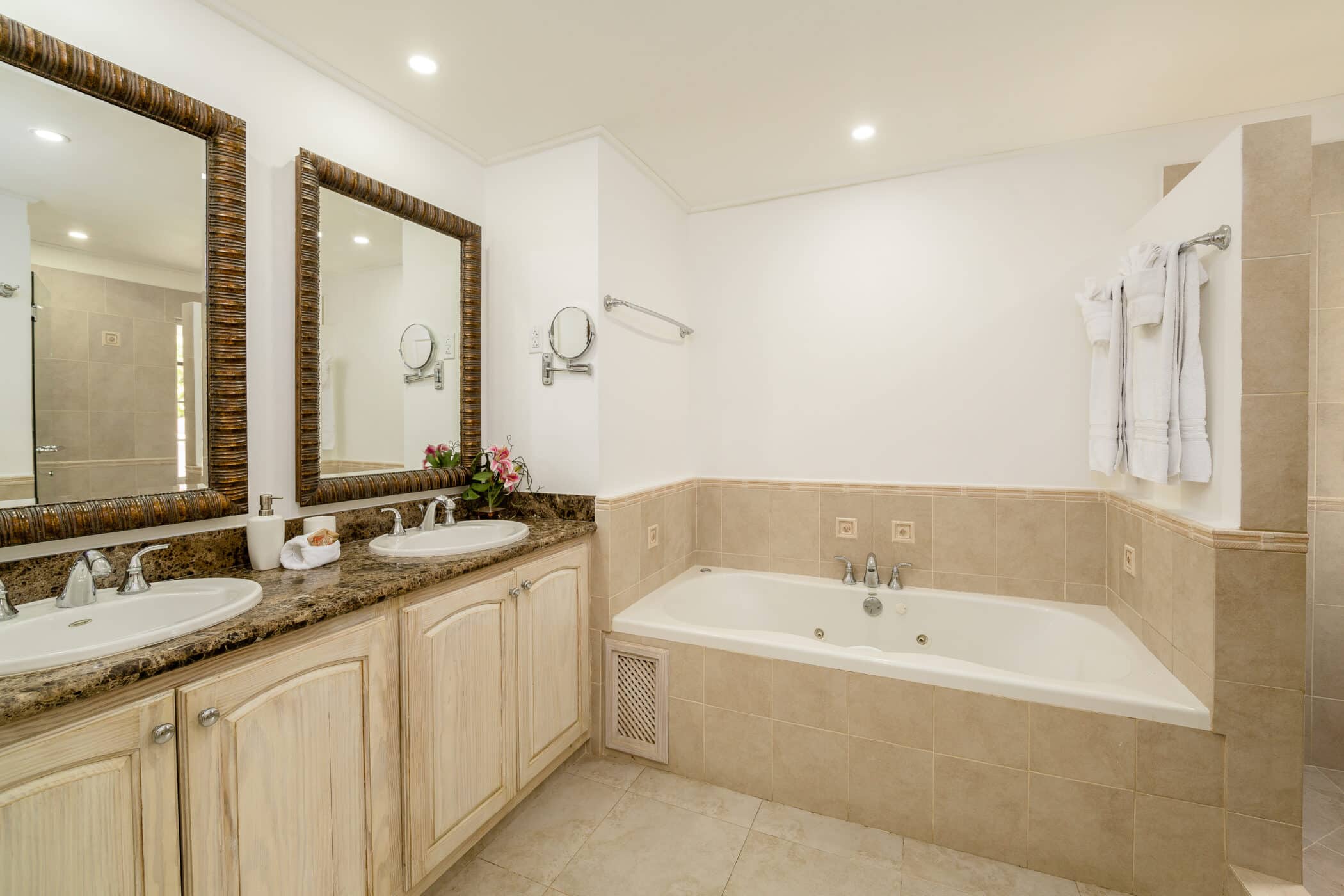 Spacious bathroom with beige tile, a double sink vanity with mirrors, a built-in bathtub, towels on a rack, and bright lighting. Flowers, including elegant Rose of Sharon blooms, and toiletries are arranged on the counter.